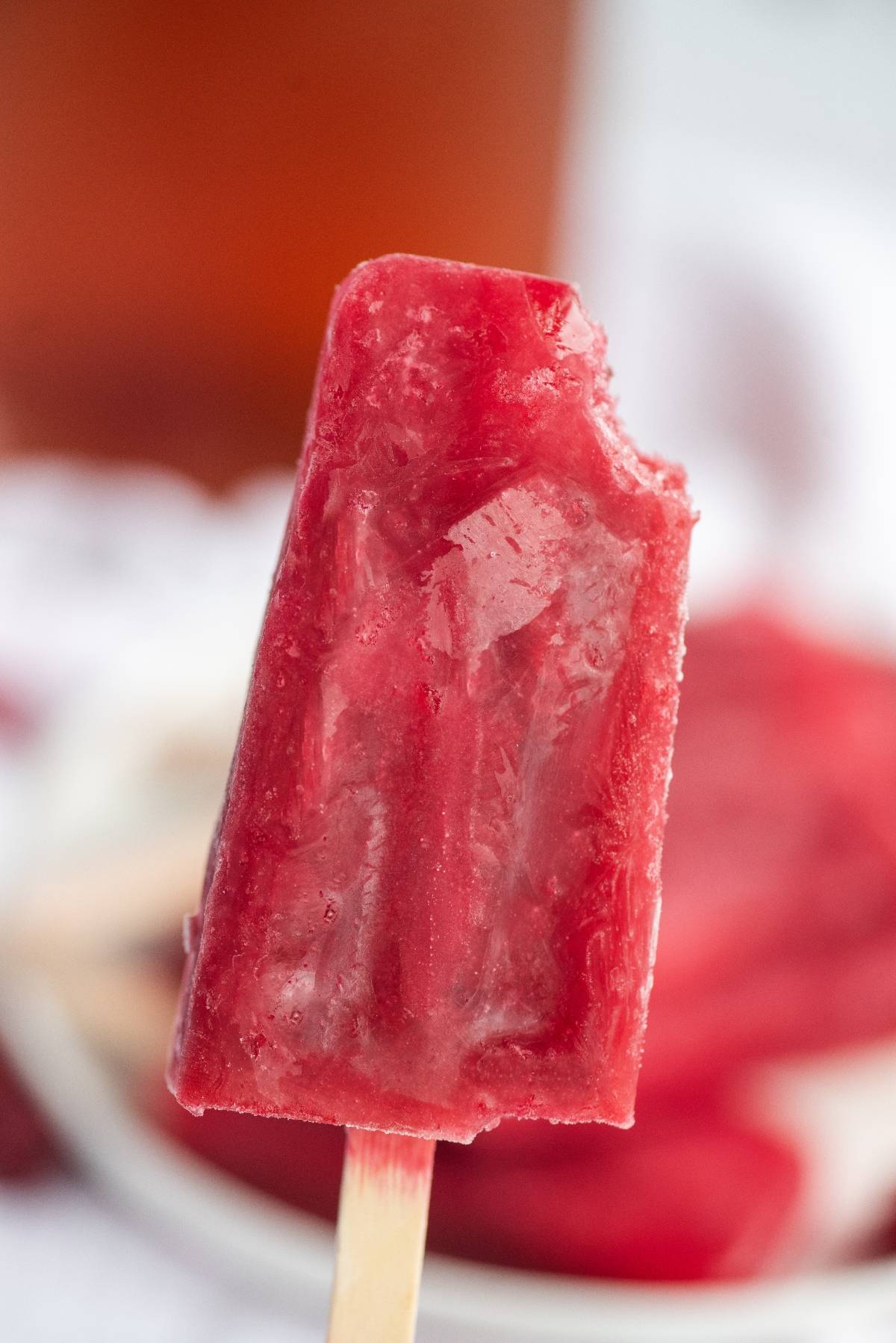 A close-up of a bitten Sweet Tea Raspberry Popsicle on a wooden stick, with more popsicles blurred in the background; perfect for summer and made using frozen or fresh raspberries.