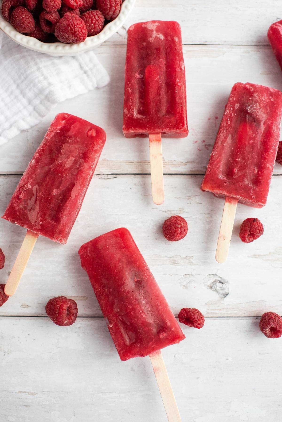 Sweet Tea Raspberry Popsicles are arranged with loose raspberries on a white wooden surface, plus a bowl of raspberries nearby—perfect for using frozen or fresh raspberries in this refreshing treat.