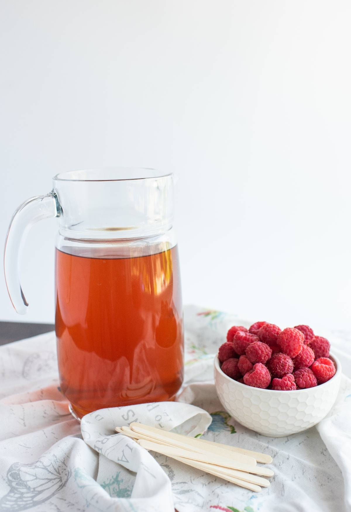 A glass pitcher of iced tea sits next to a bowl of fresh raspberries and wooden sticks on a white cloth, perfect for making Sweet Tea Raspberry Popsicles.