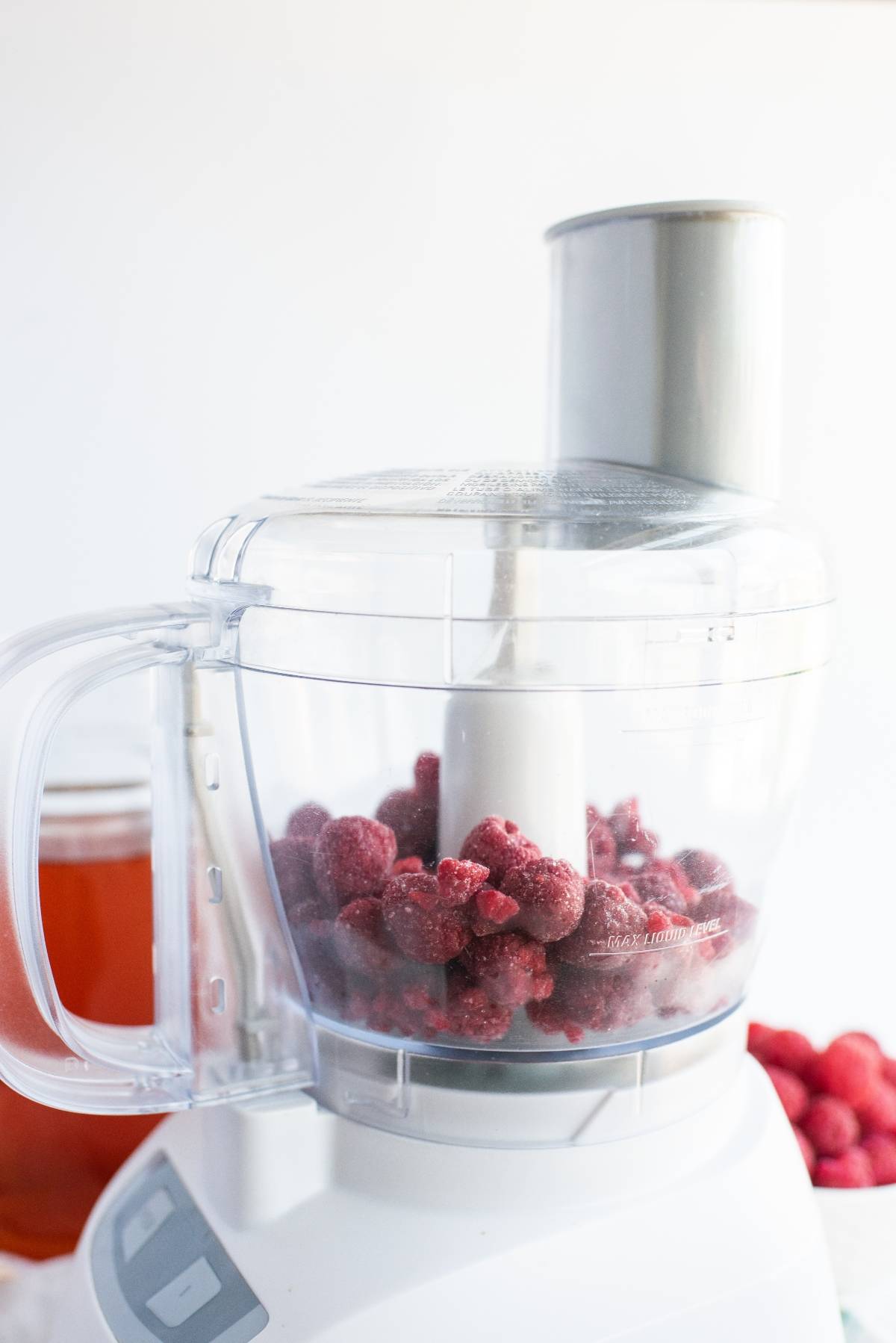A food processor filled with frozen raspberries, perfect for a raspberry ice pop recipe, with a jar and more berries in the background.