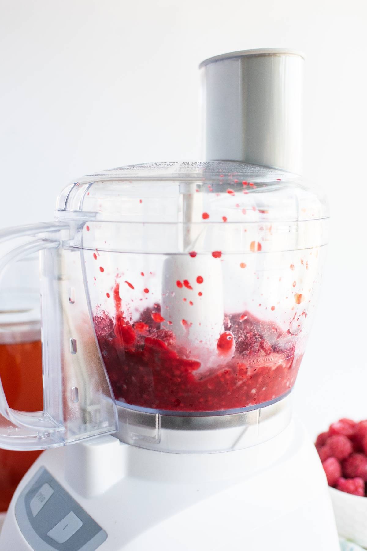 Food processor blending red raspberries for a sweet tea raspberry popsicles mix, with splatters inside the container on a white background.