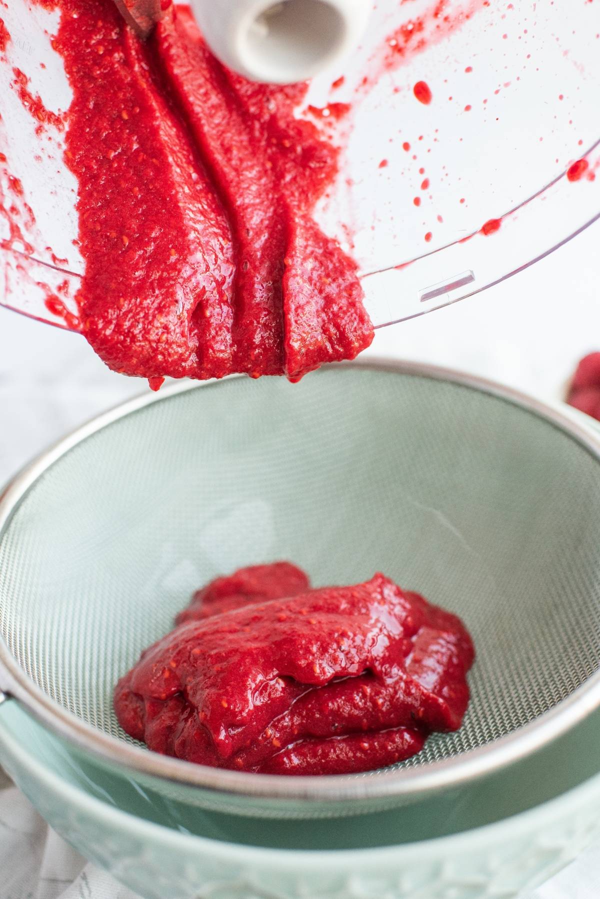 Bright red raspberry puree being poured from a blender into a fine mesh strainer over a bowl, perfect for crafting Sweet Tea Raspberry Popsicles or any sweet summer treats.
