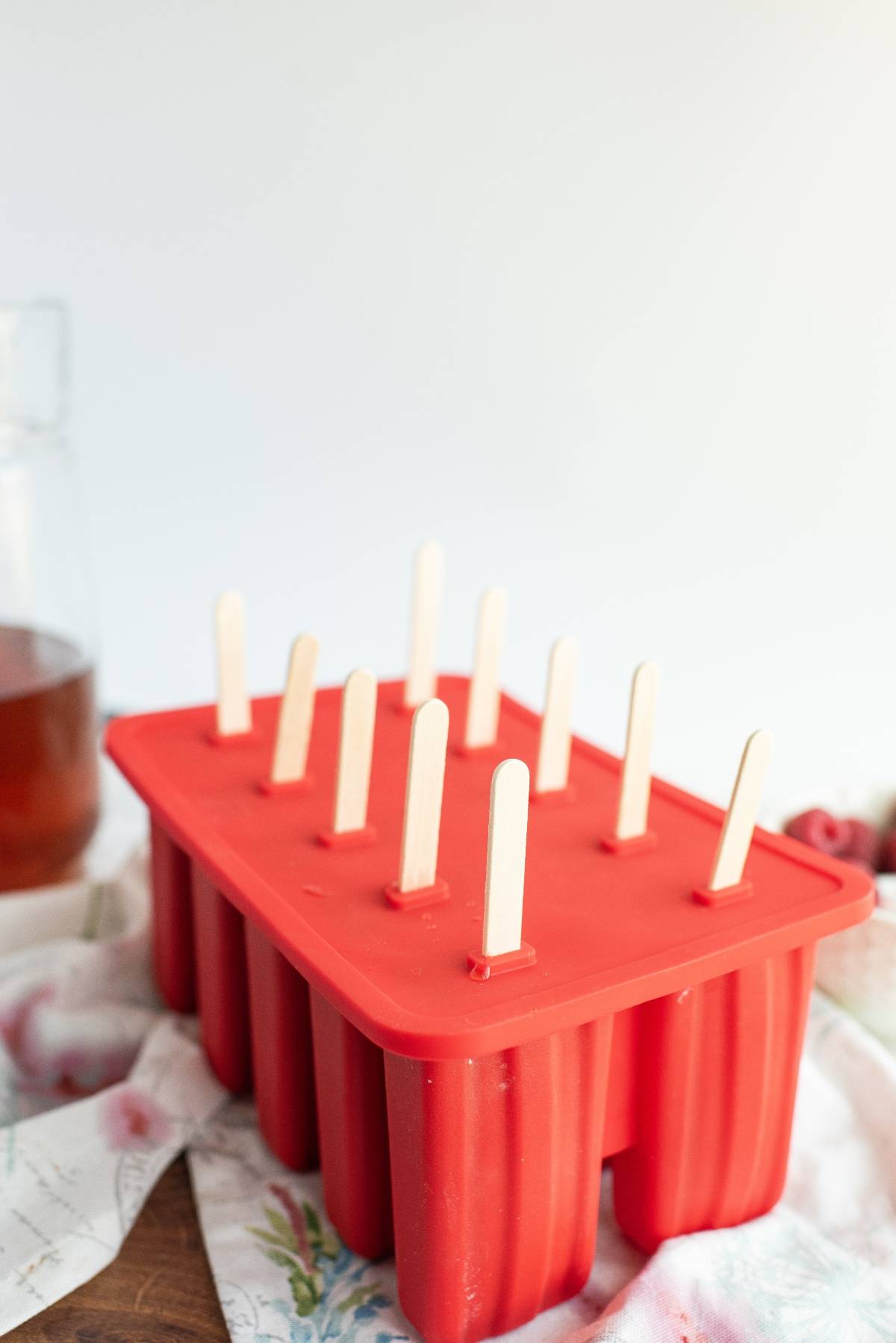 Red popsicle mold with wooden sticks inserted, sitting on a patterned cloth—perfect for sweet summer treats like raspberry ice pop recipes or popsicles made with sweetened ice tea.