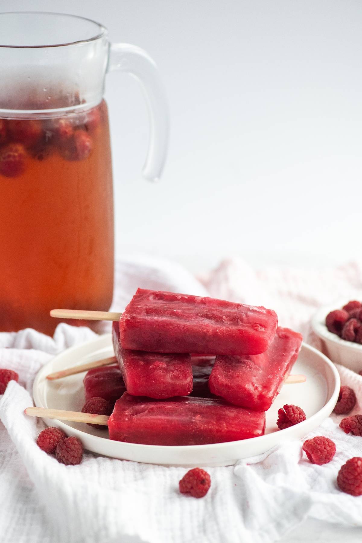 A plate of Sweet Tea Raspberry Popsicles with raspberries, next to a pitcher of iced raspberry tea and a bowl of fresh raspberries—perfect treats made using frozen or fresh raspberries.