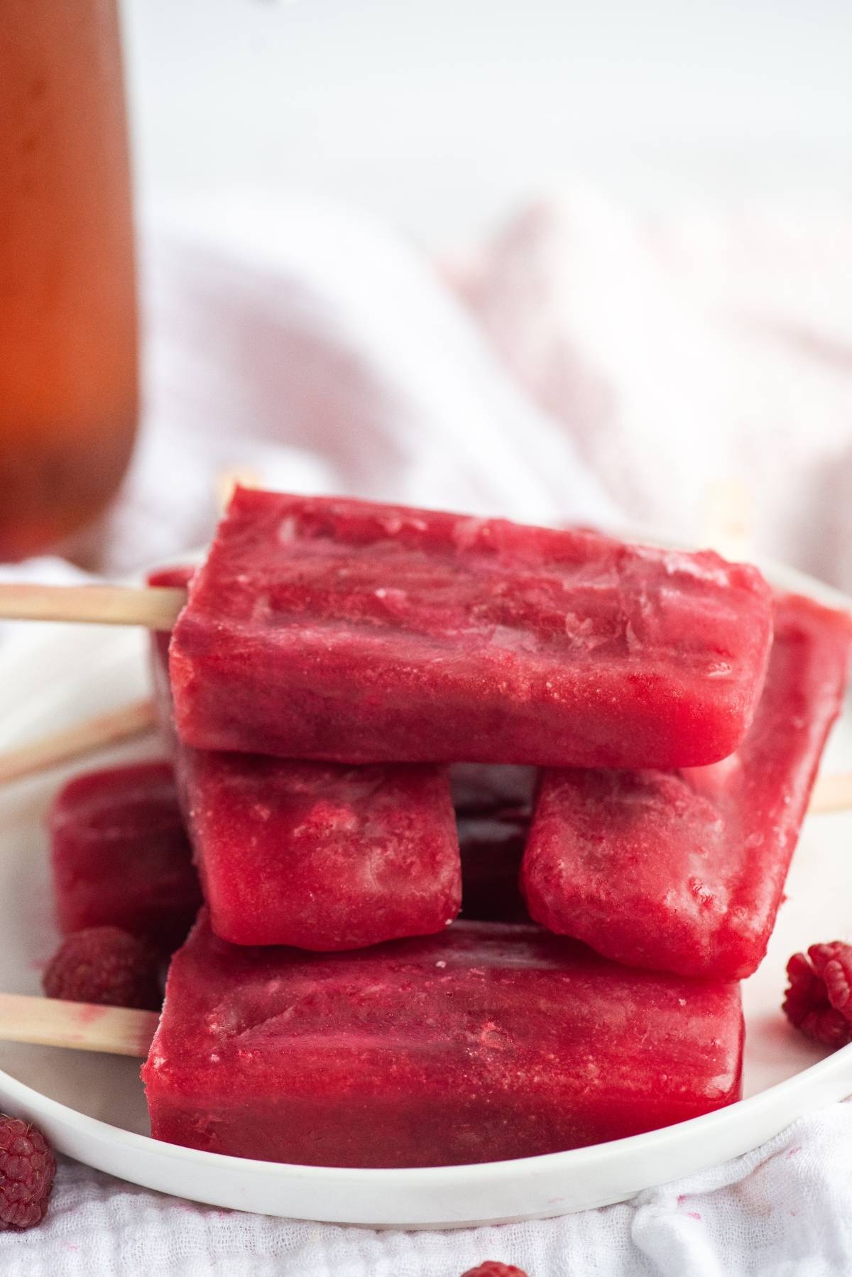 Four bright red raspberry popsicles made with sweetened ice tea are stacked on a white plate, with a few raspberries beside them—perfect sweet summer treats.