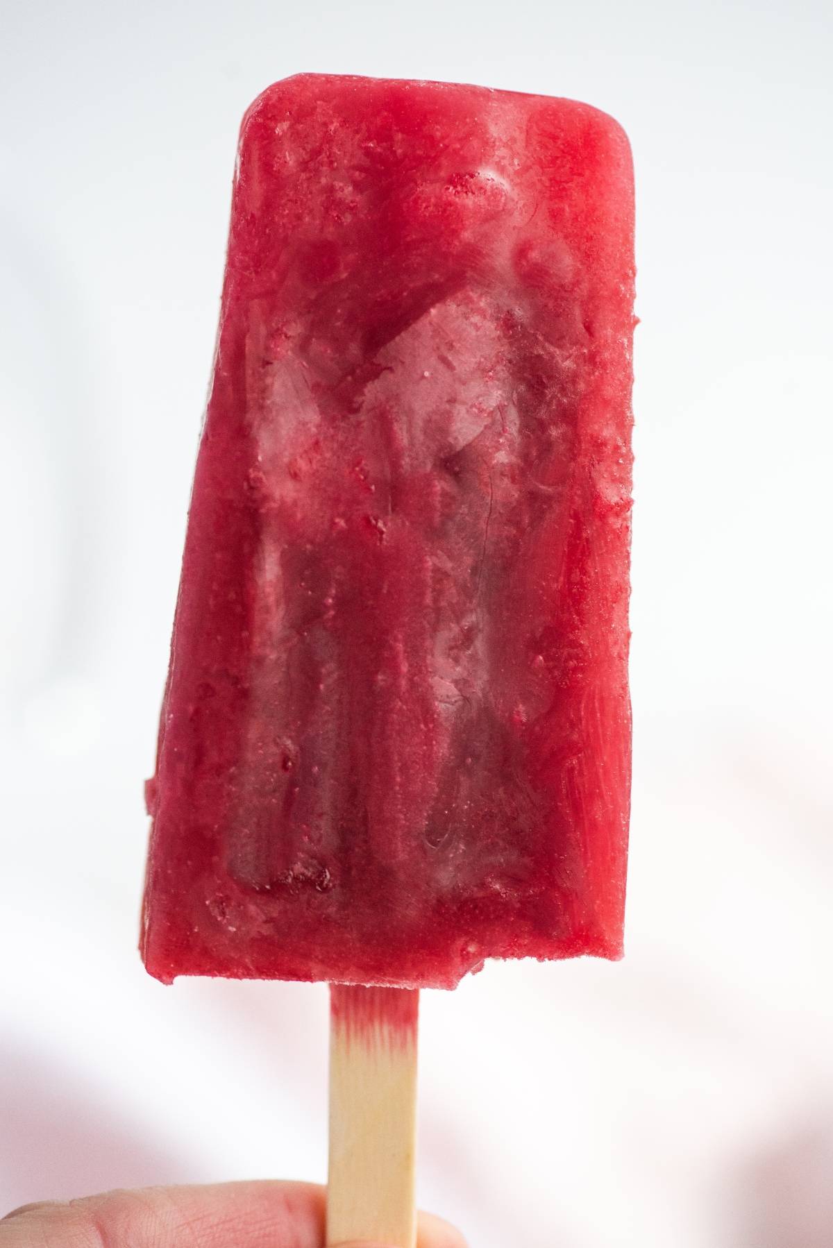 A hand holding a bright red popsicle on a wooden stick against a white background, perfect for those craving sweet summer treats.