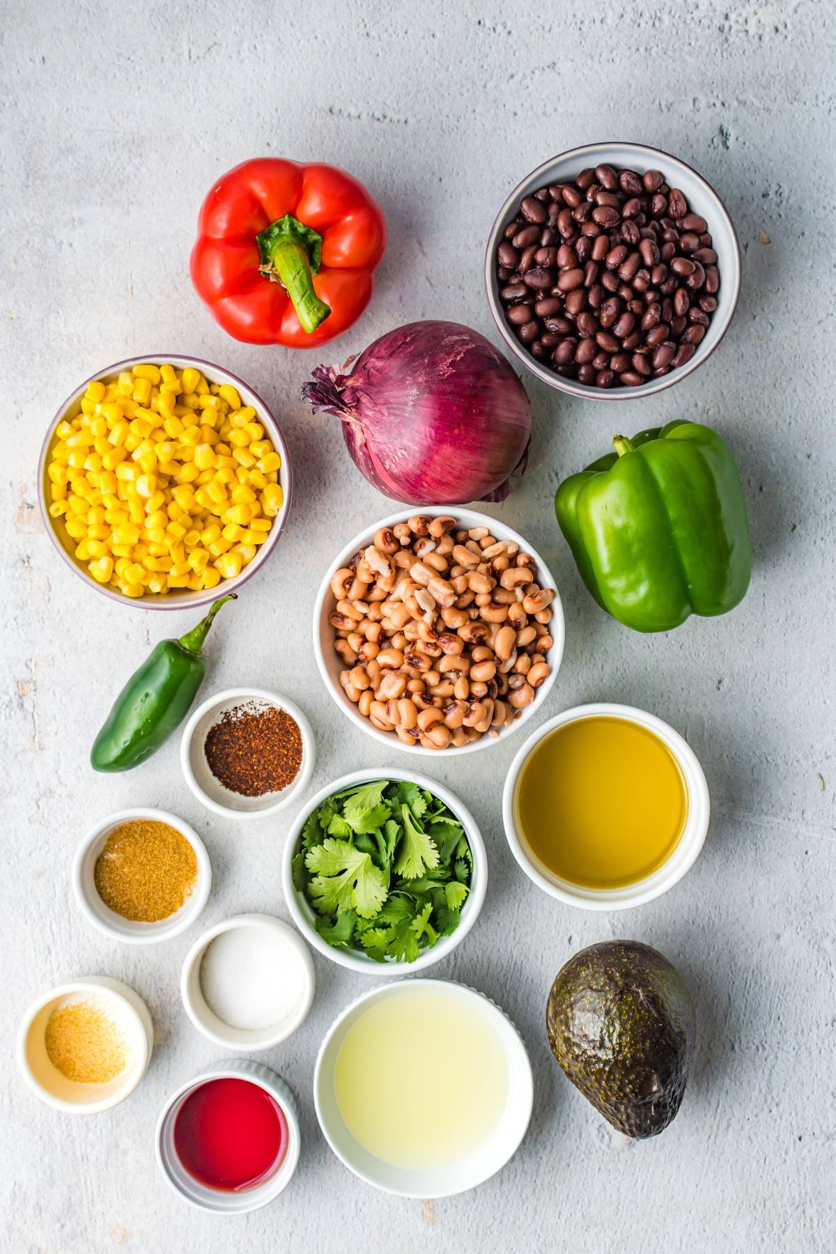 Assorted fresh vegetables, beans, herbs, and spices arranged in bowls on a light gray surface—perfect ingredients for a Texas Cowboy Caviar dip or a flavorful Southwest vegan dip at your next summer BBQ.