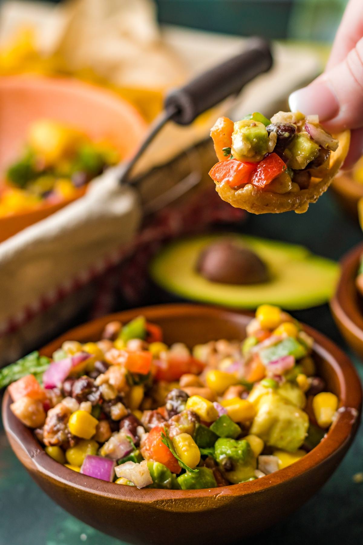 A hand holds a tortilla chip with vibrant bean and corn salsa, a delicious Southwest vegan dip; a bowl of this healthy summer BBQ dip sits below.