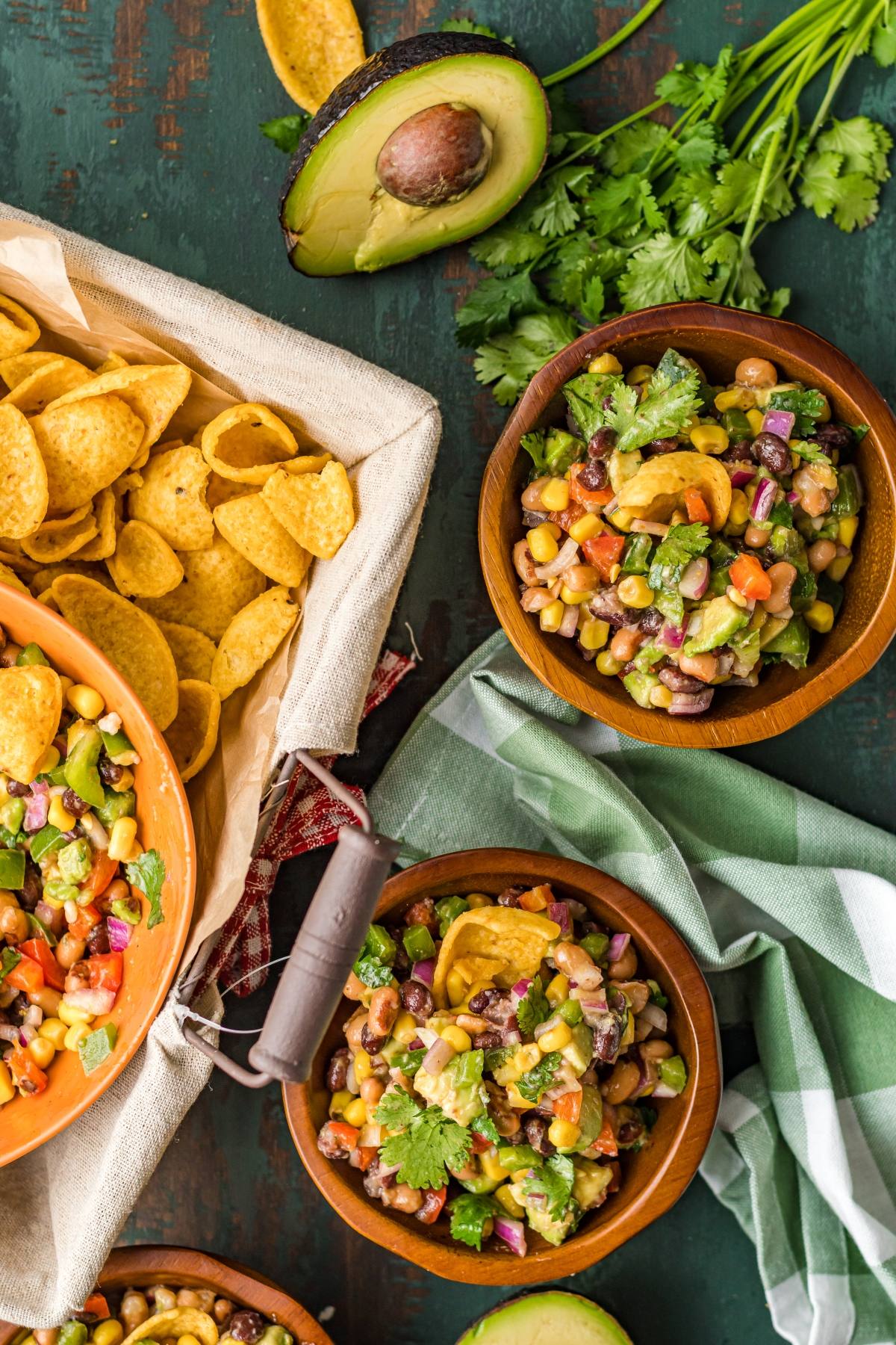 Bowls of colorful Texas Cowboy Caviar dip—an irresistible Southwest vegan dip—sit beside tortilla chips, fresh cilantro, and halved avocado on a table.