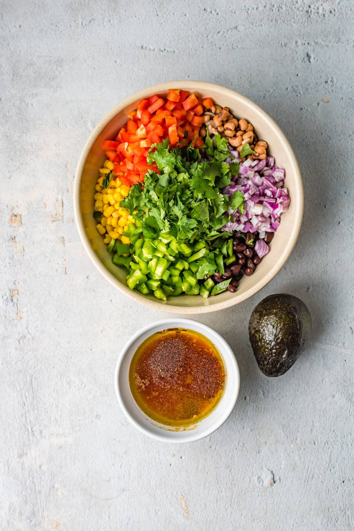 A bowl with chopped veggies and beans, a smaller bowl of dressing, and a whole avocado on a gray surface—perfect for serving as a Southwest vegan dip at your next summer gathering.