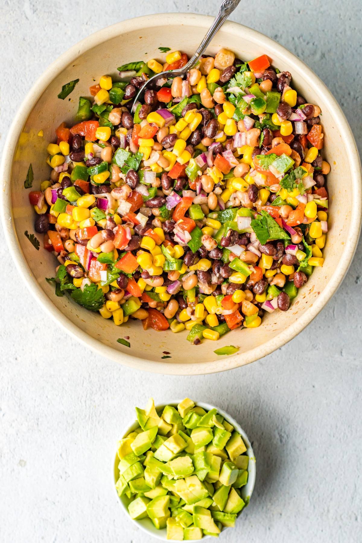 A bowl of colorful bean and corn salsa featuring tomatoes and herbs, sits next to a small bowl of diced avocado.
