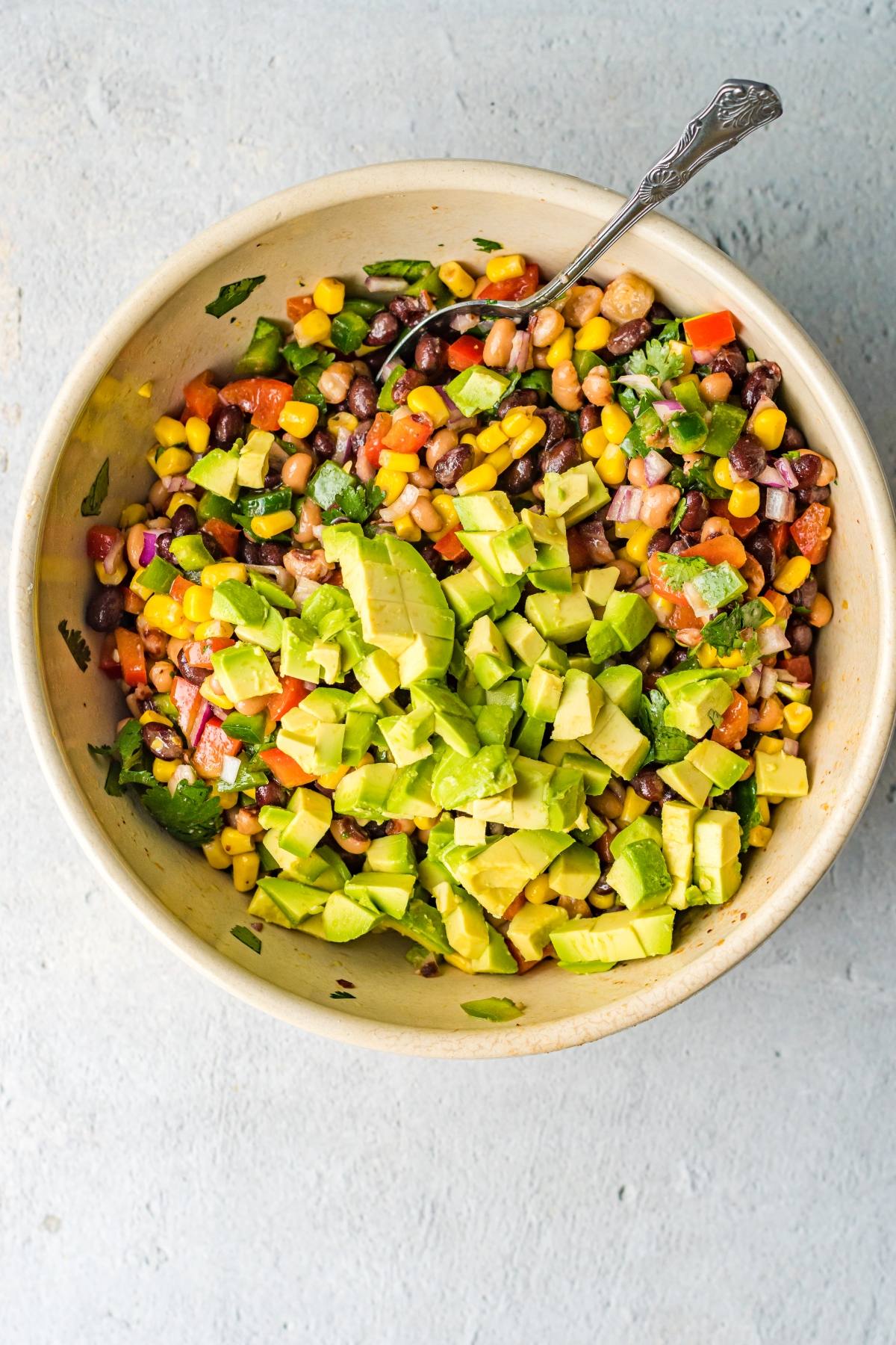 A bowl of chopped avocado, corn, beans, tomatoes, and vegetables with a spoon on a light background—perfect for serving as a fresh Bean and corn salsa or classic Texas Cowboy Caviar dip.