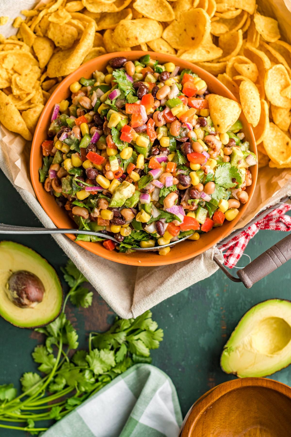 A bowl of colorful Texas Cowboy Caviar dip with corn chips, surrounded by avocado halves and fresh cilantro.