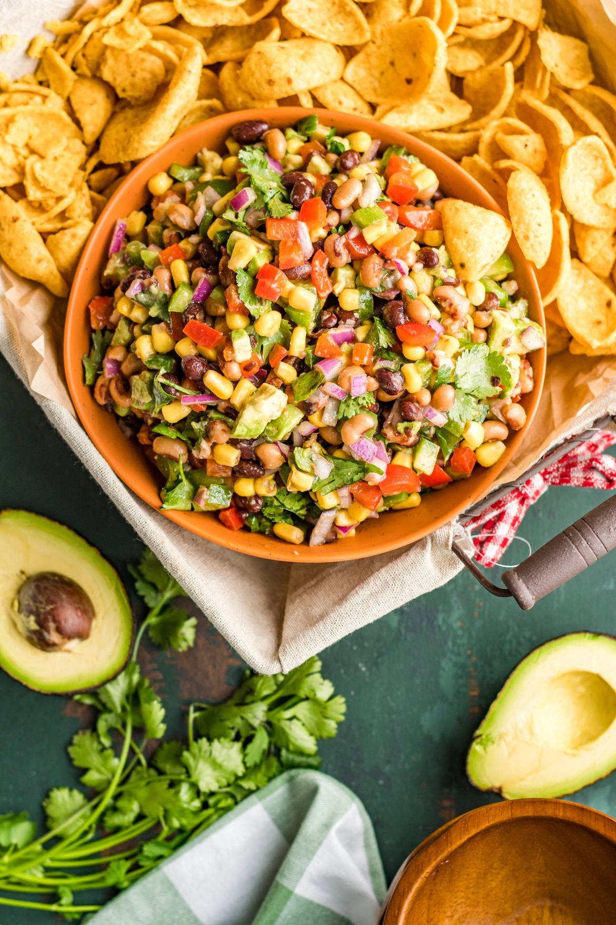 A bowl of colorful bean and corn salsa, a Southwest vegan dip, surrounded by tortilla chips, avocado, and fresh cilantro—perfect as a healthy summer BBQ dip or colourful appetizer.