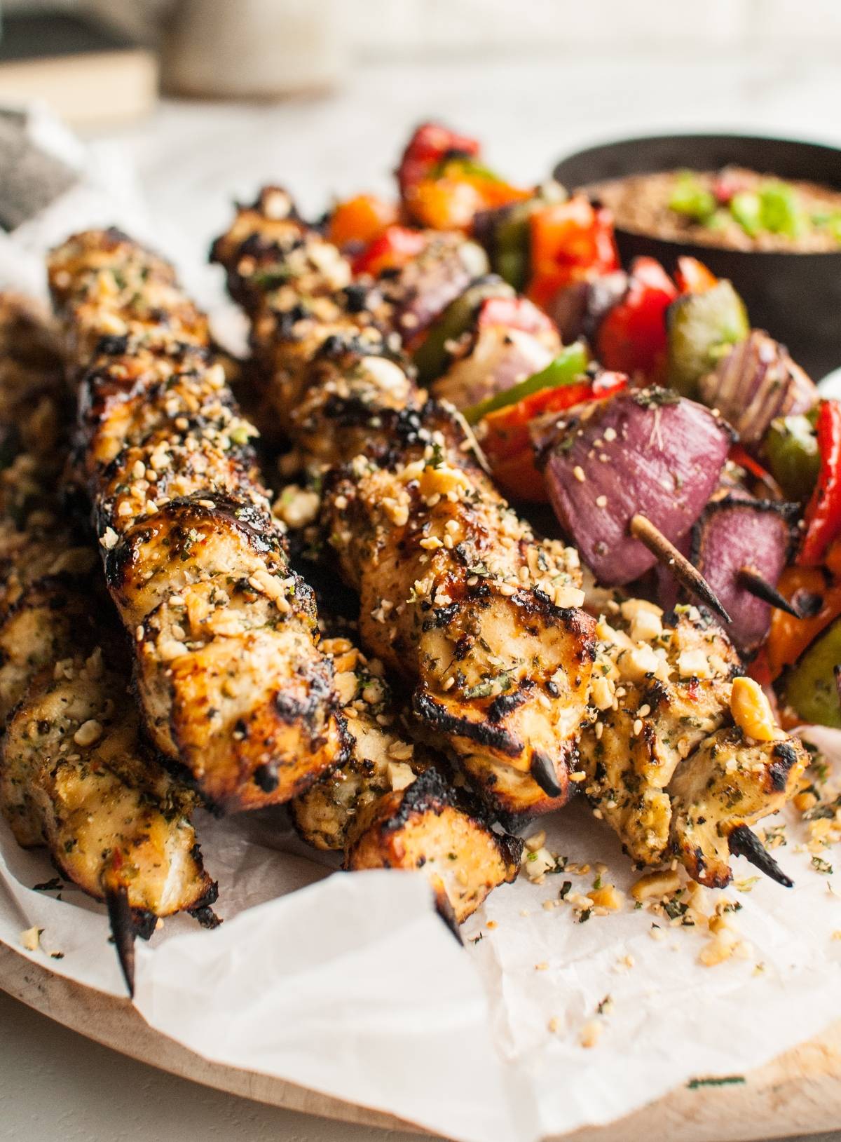 Grilled Thai chicken skewers with herbs and chopped nuts, served alongside grilled vegetables on parchment paper, paired with a creamy cashew dipping sauce.