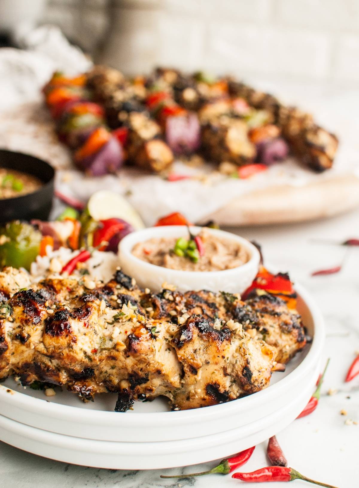 Thai Spicy Basil Chicken Skewers served on a plate with rice, vegetables, and a flavorful cashew dipping sauce; more skewers in the background.