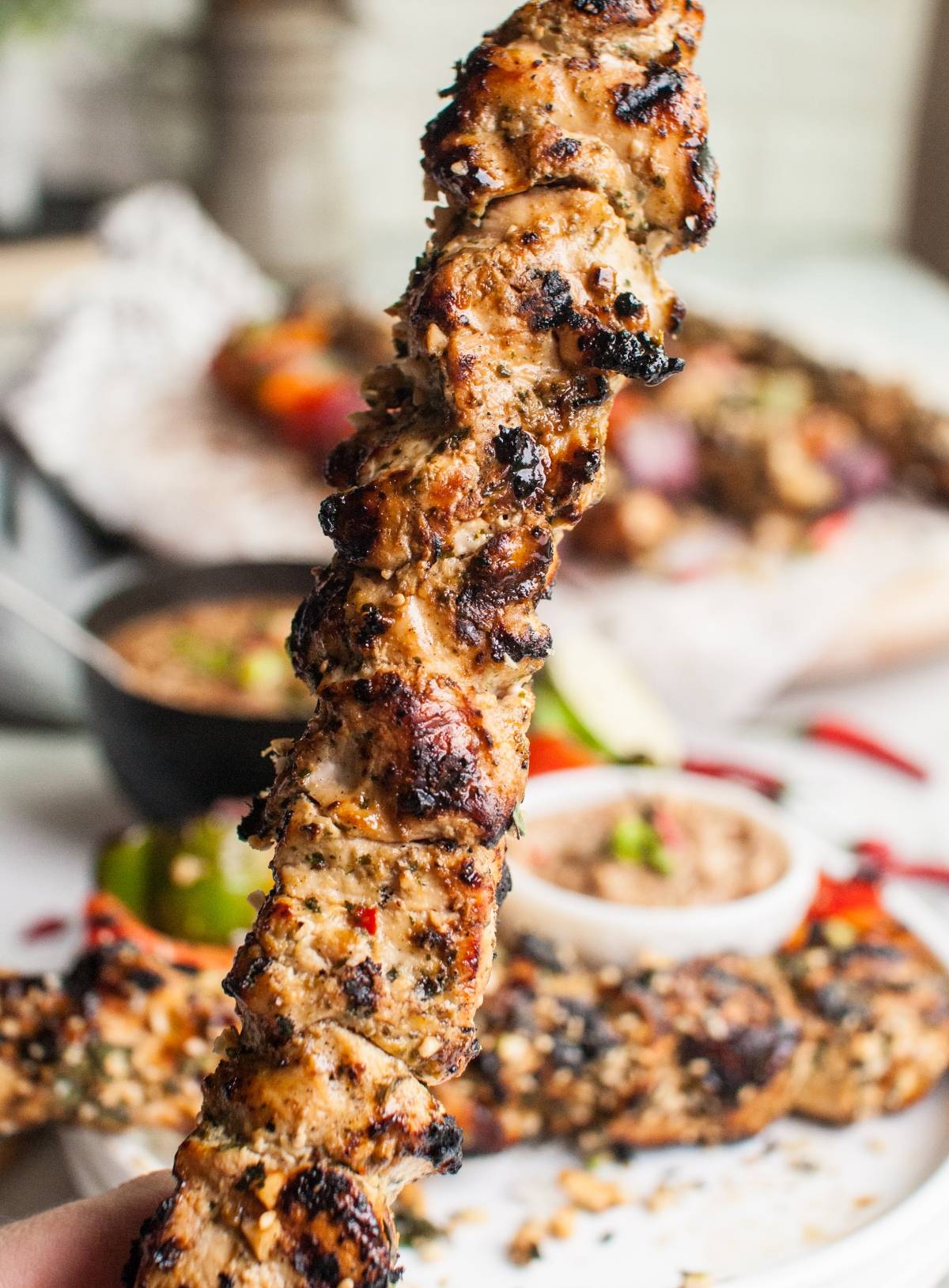 Close-up of a grilled chicken skewer with Dairy free skewer marinade held up, Cashew dipping sauce and more skewers blurred in the background.