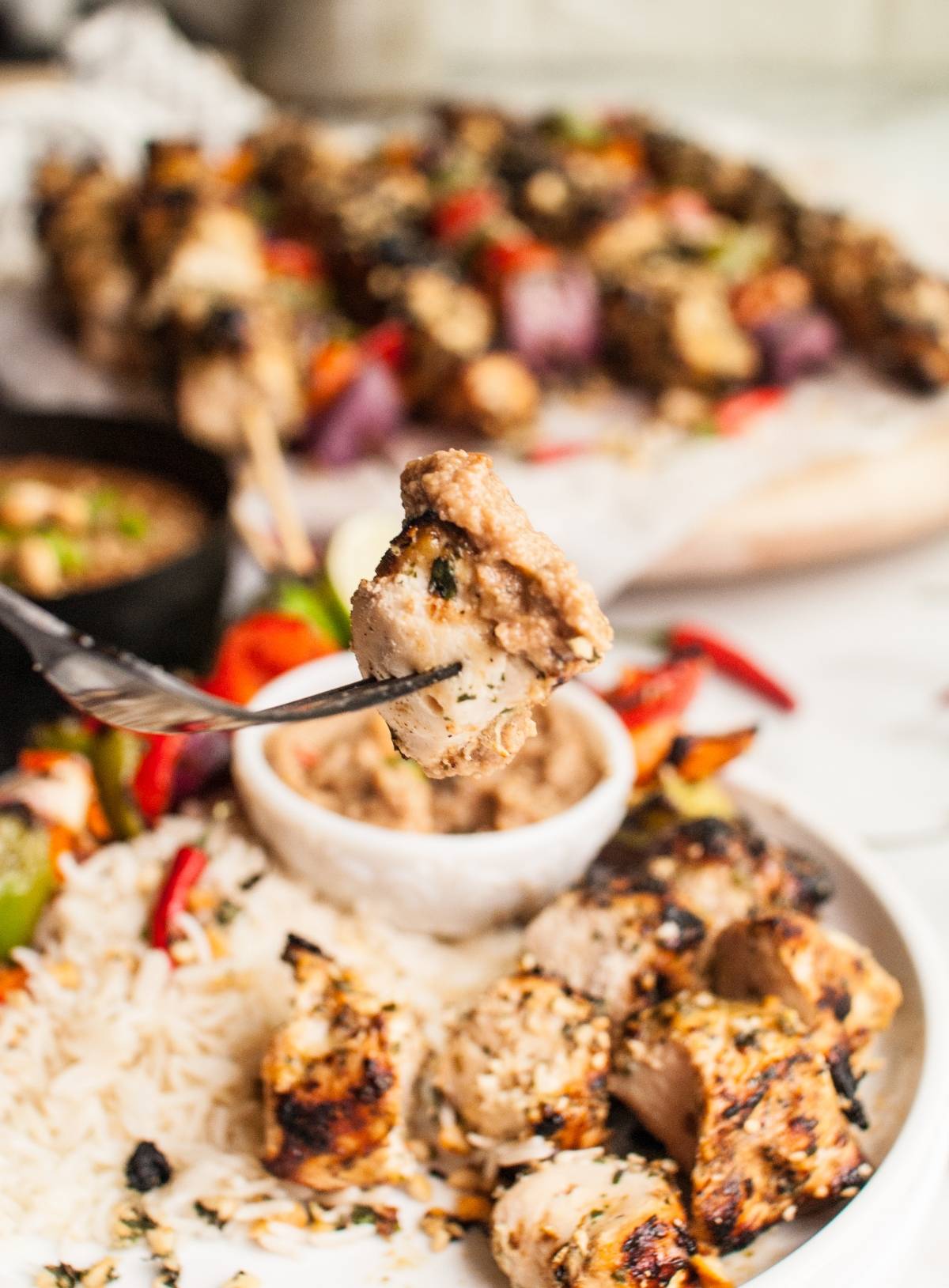 A fork holds a piece of Thai Spicy Basil Chicken Skewer above a plate with rice, vegetables, and cashew dipping sauce.