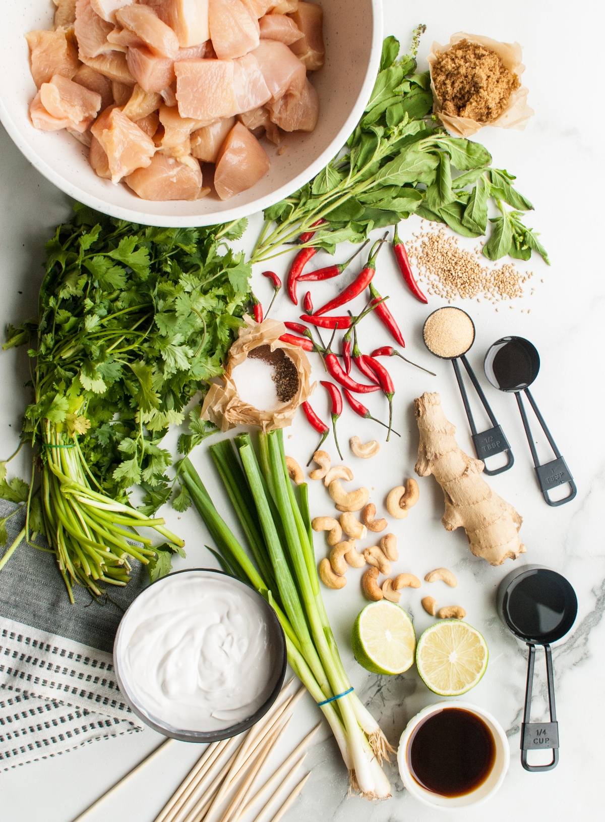 Raw chicken, fresh herbs, chilies, ginger, limes, sauces, brown sugar, and measuring spoons on a white surface—perfect for making Thai chicken skewers with vibrant flavors and aromatic spices.