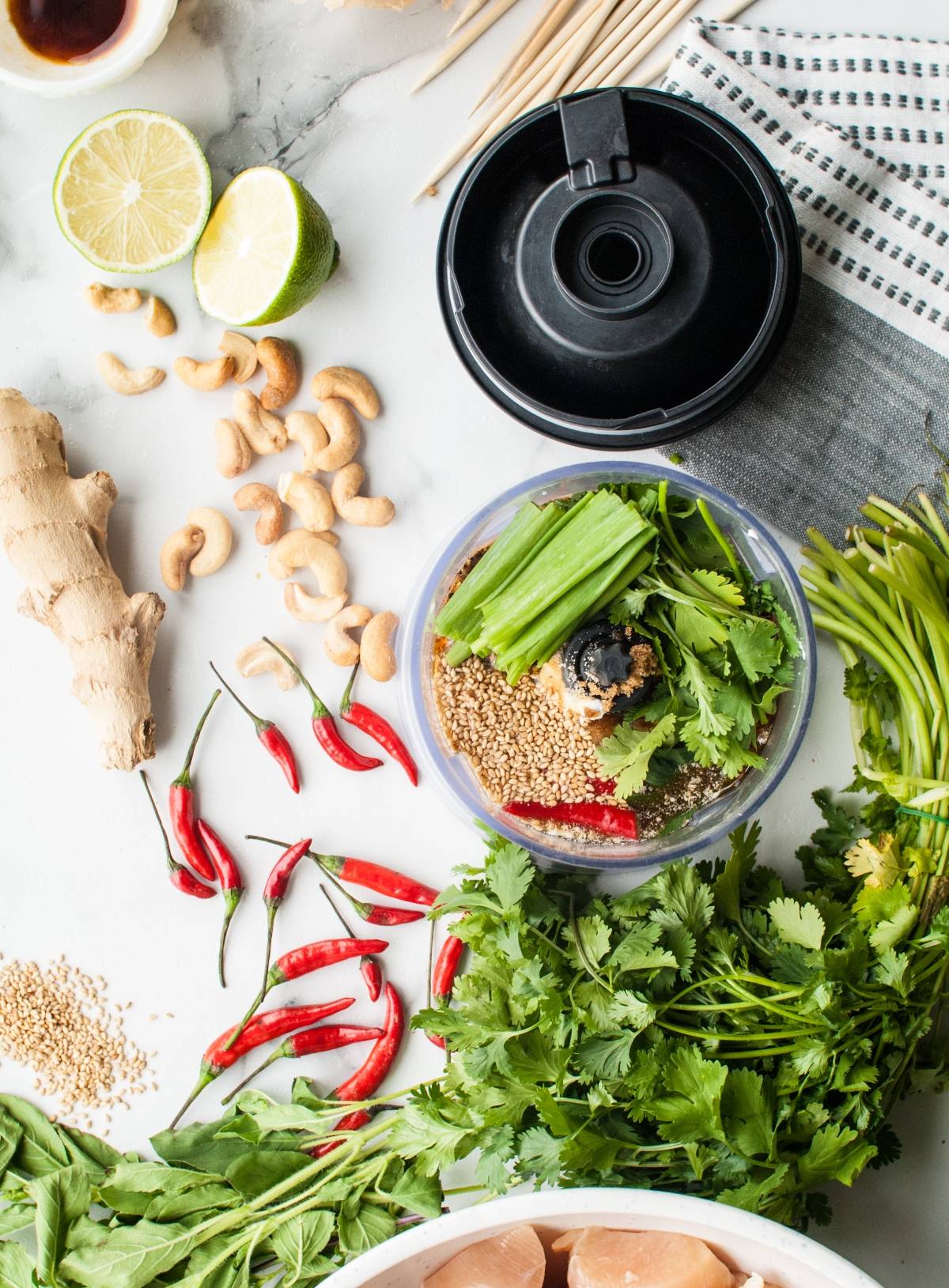 Top view of fresh herbs, chilies, cashews, ginger, and other ingredients for Thai chicken skewers or kebabs arranged around a blender cup on a white surface.
