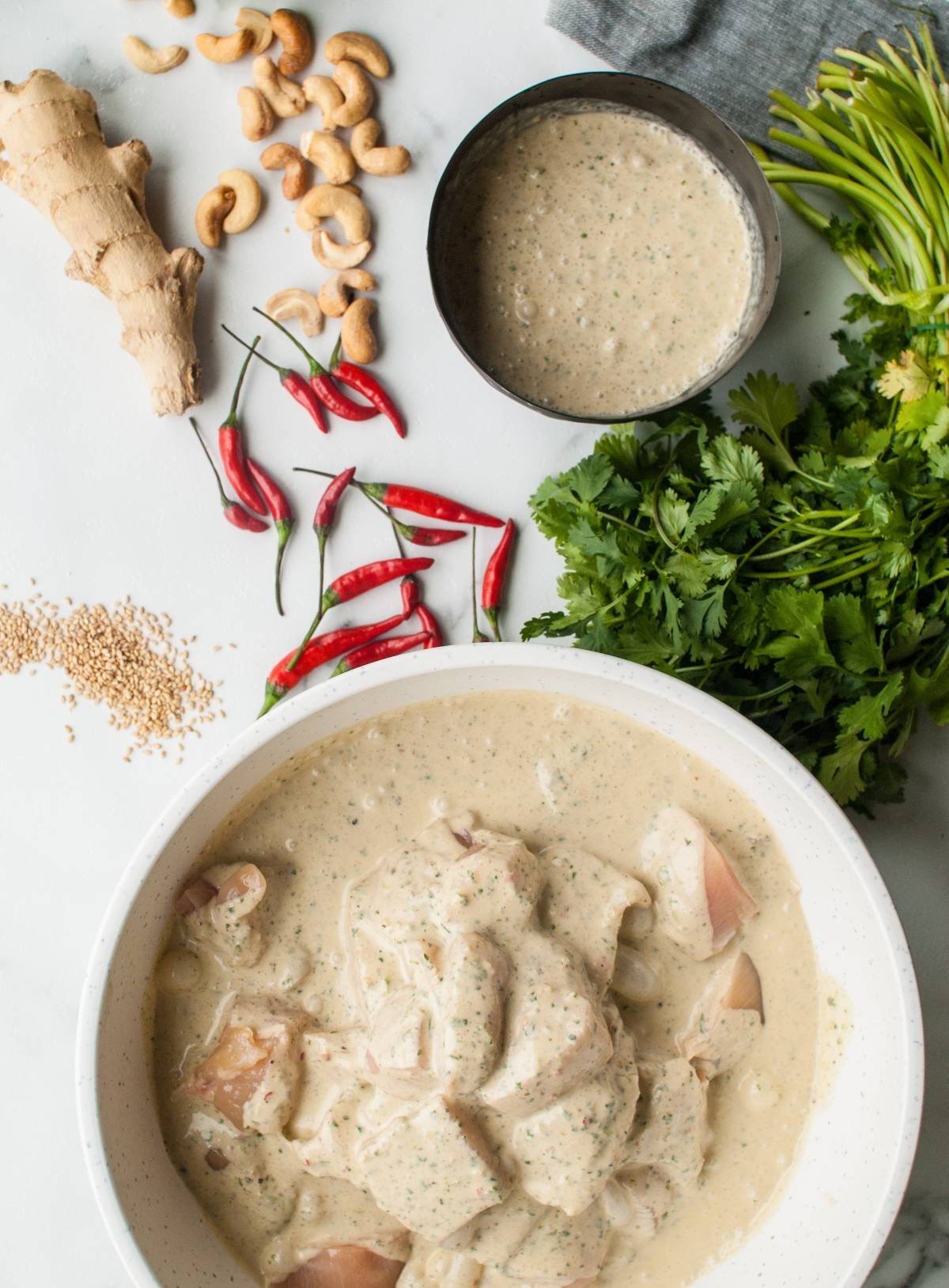 Bowl of marinating chicken in creamy sauce, surrounded by herbs, ginger, chilies, cashews, and sesame seeds—perfect for making Thai Spicy Basil Chicken Skewers or kebabs.