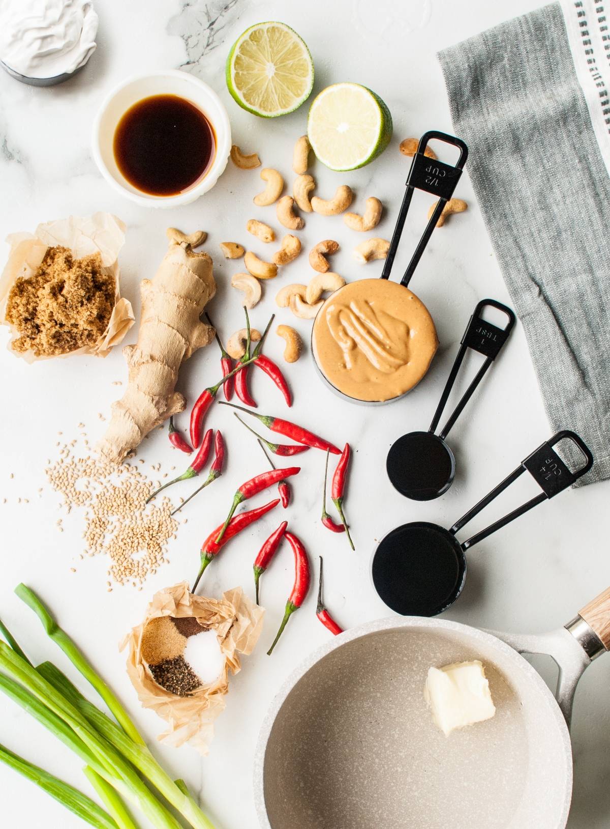 Overhead view of peanut sauce ingredients—peanut butter, ginger, lime, chilies, cashews, soy sauce, and measuring cups—perfect for pairing with Thai chicken skewers or kebabs.