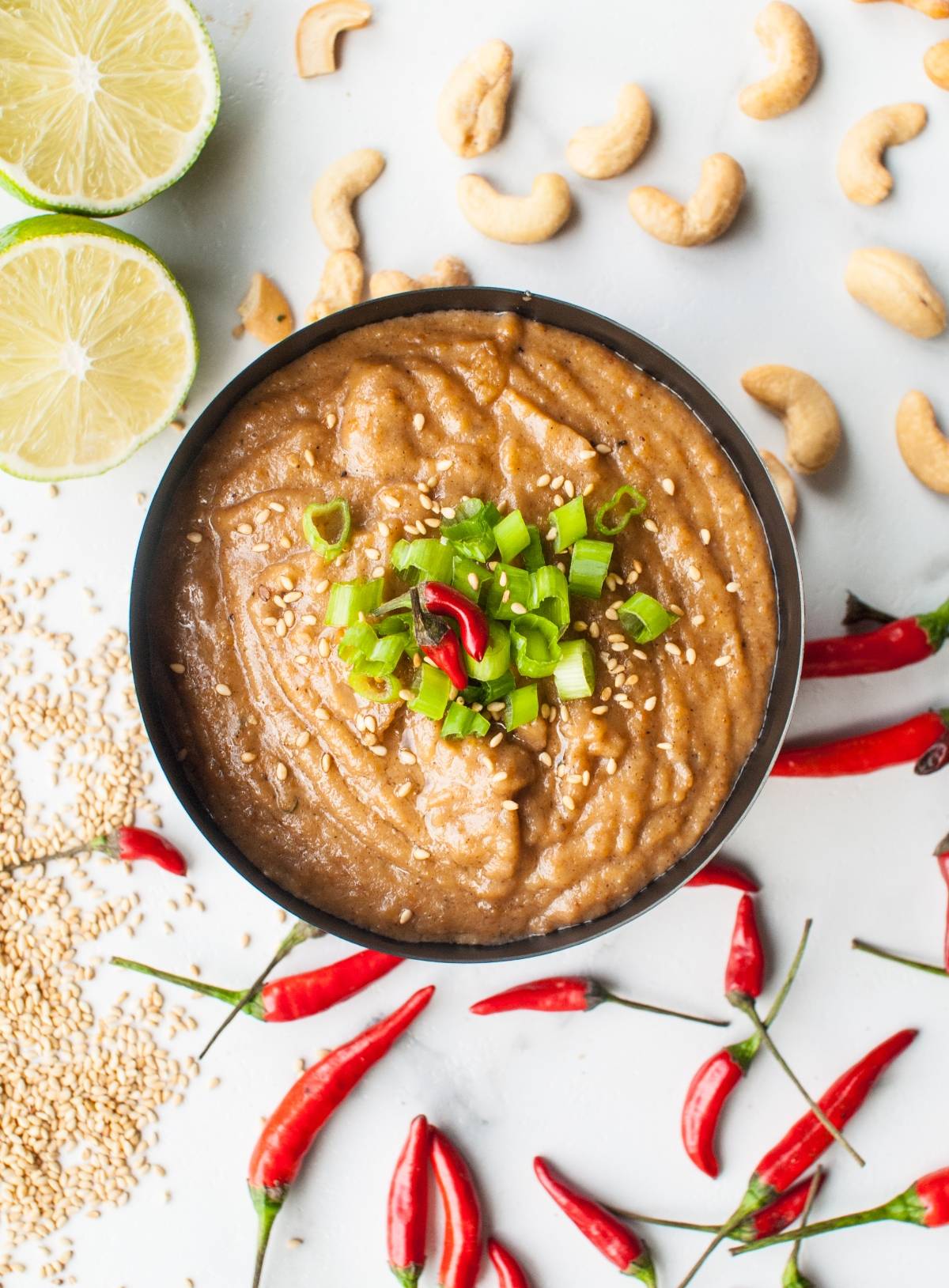 A bowl of creamy cashew dipping sauce topped with chopped green onions and chili, surrounded by limes, cashews, and red chilies—perfect for pairing with Thai chicken skewers or kebabs.