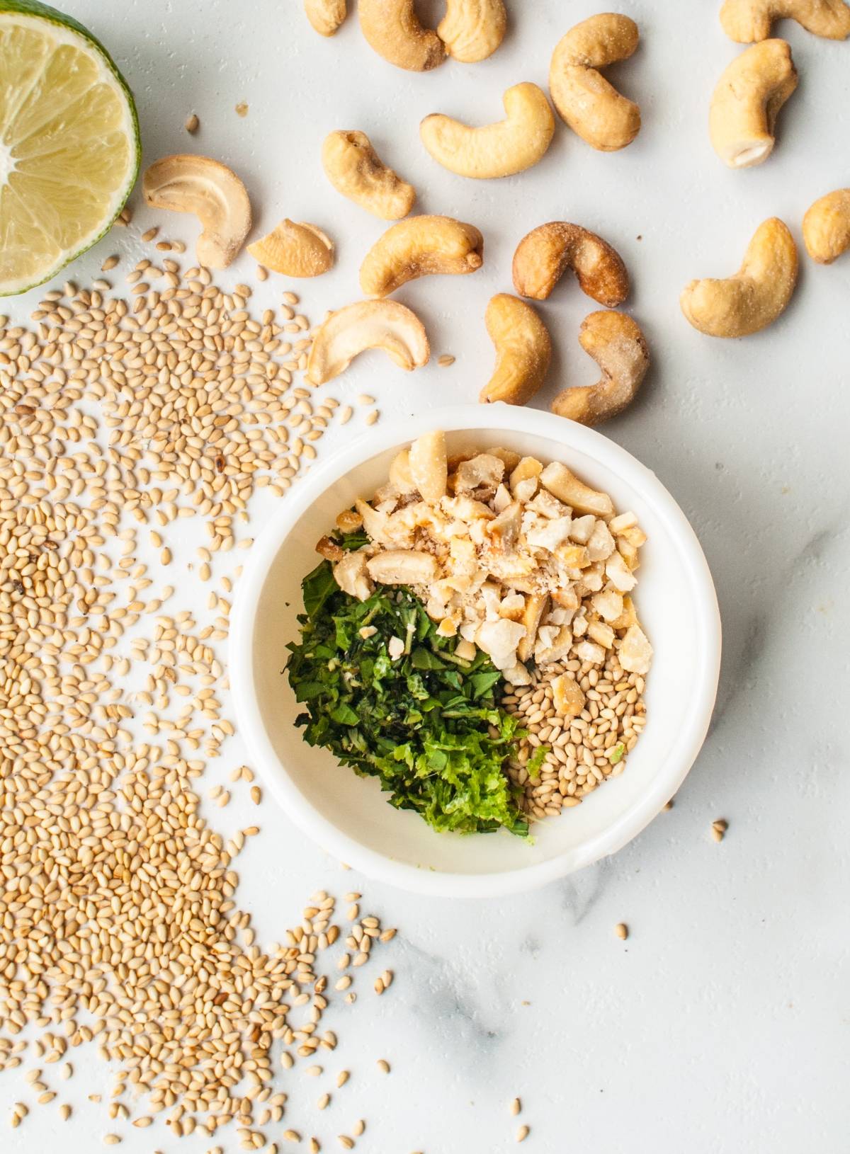 A bowl with chopped cashews, cilantro, and sesame seeds—perfect as a Cashew dipping sauce or Gremolata Topping—surrounded by more cashews, sesame seeds, and lime.