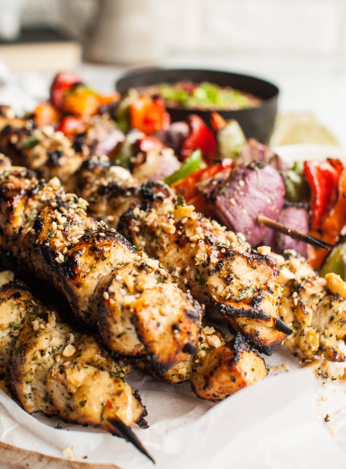 Thai Spicy Basil Chicken Skewers and vegetables are grilled to perfection, served on parchment paper with a bowl of cashew dipping sauce in the background.