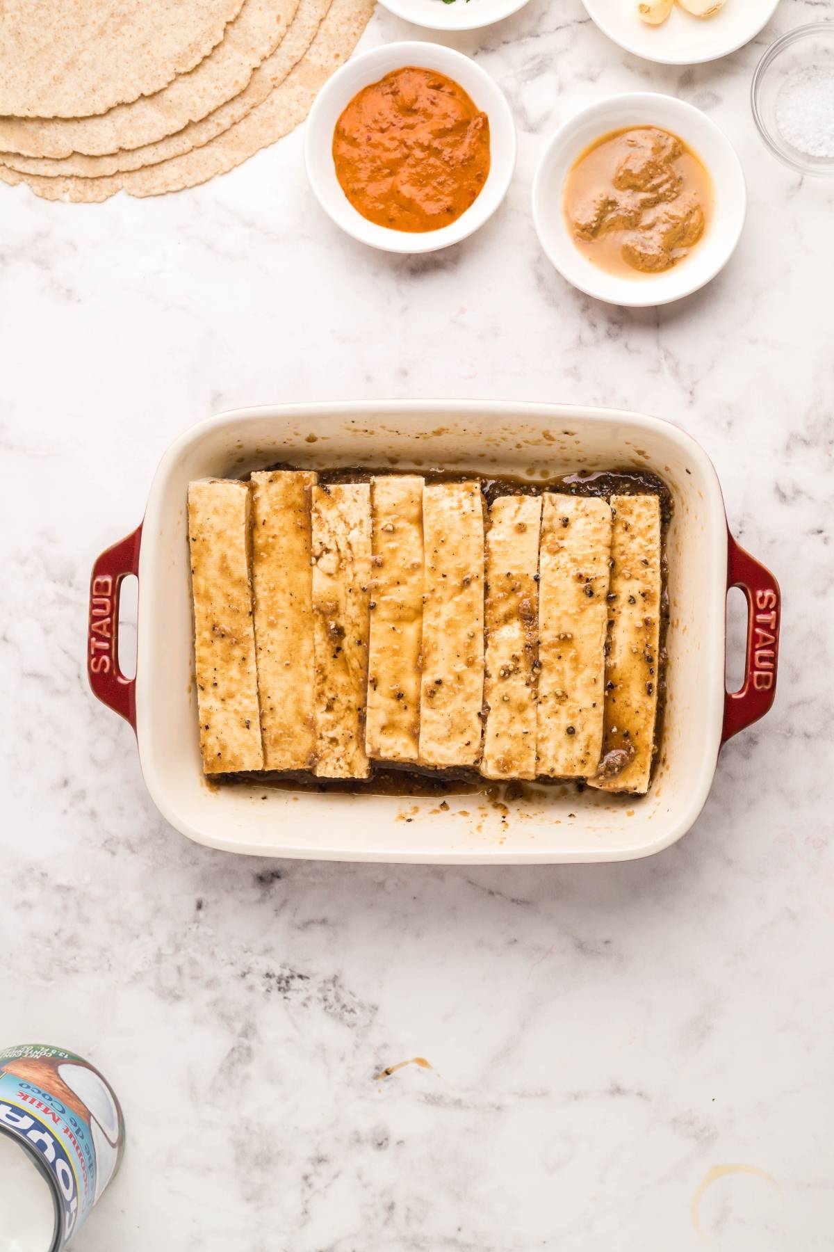 Sliced tofu marinating in a white Staub dish, surrounded by sauces like Coconut Almond Butter Dip and tortillas—perfect for assembling an easy tofu wrap that's both vegan and gluten free, all set on a marble countertop.