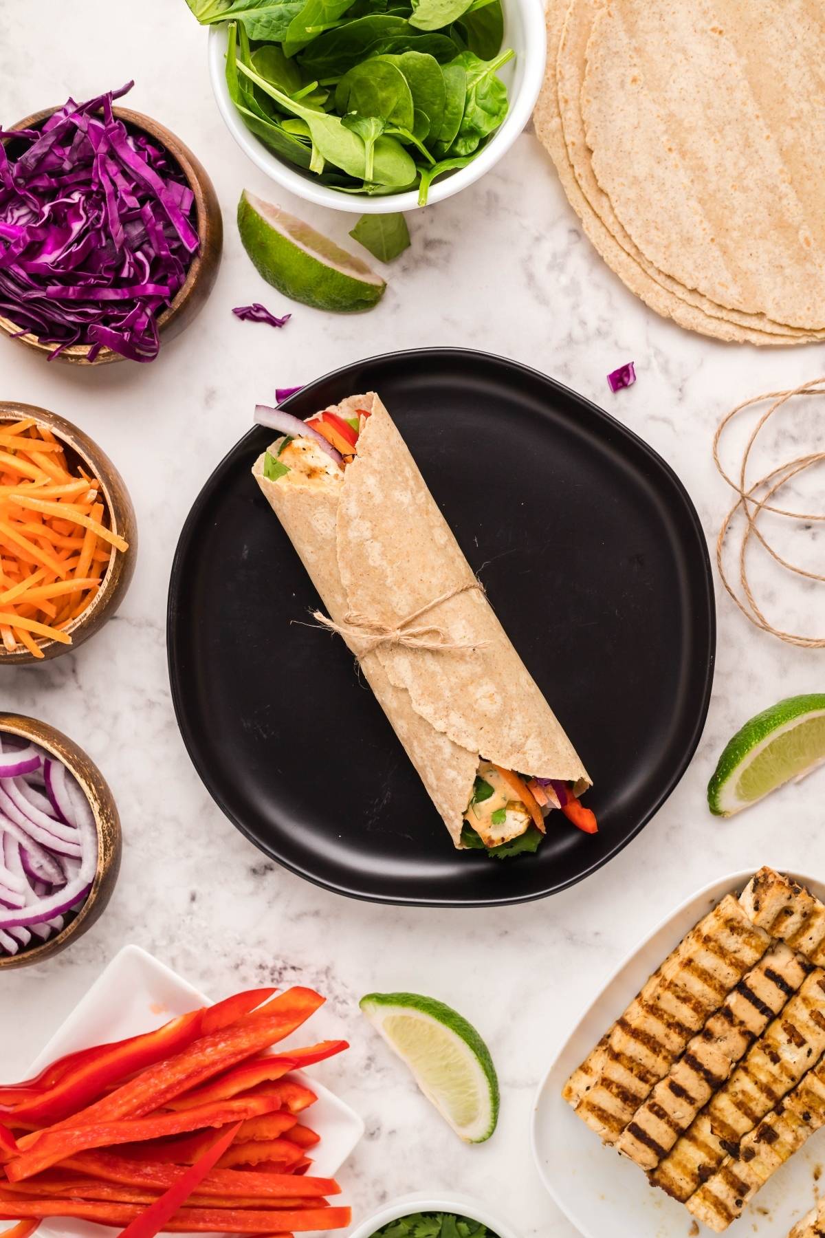 A wrapped tortilla filled with veggies sits on a black plate, surrounded by bowls of fresh chopped vegetables—a delicious Tofu Thai Wrap that's perfect for a vegan and gluten free lunch.