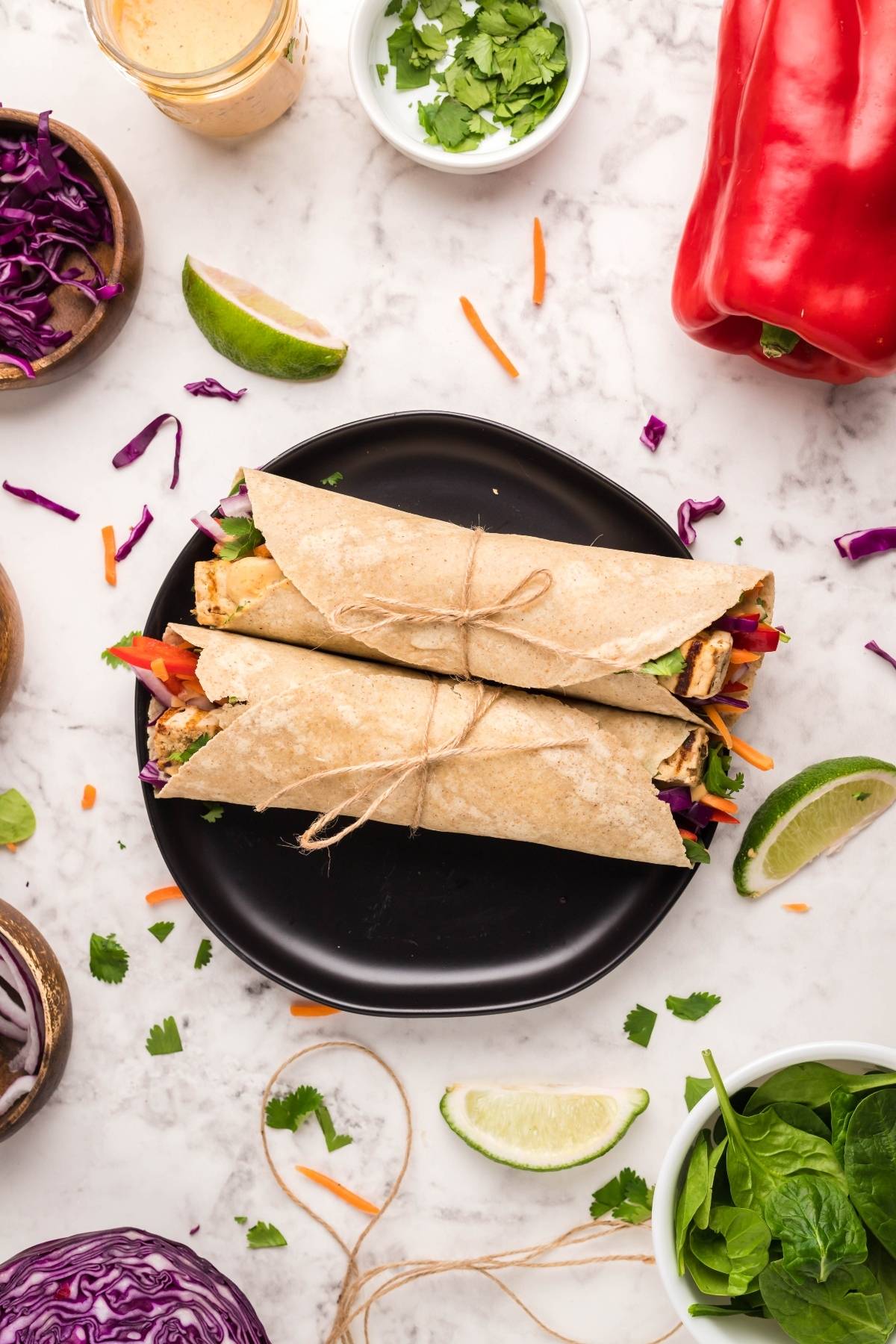 Two veggie wraps tied with twine on a black plate, surrounded by fresh vegetables and lime wedges—an easy tofu wrap perfect for a vegan gluten free lunch.