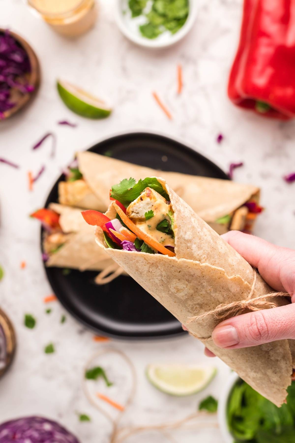 Hand holding a Tofu Thai Wrap filled with carrots, red cabbage, and cilantro—an ideal Vegan and gluten free wrap—with more wraps arranged on a plate.