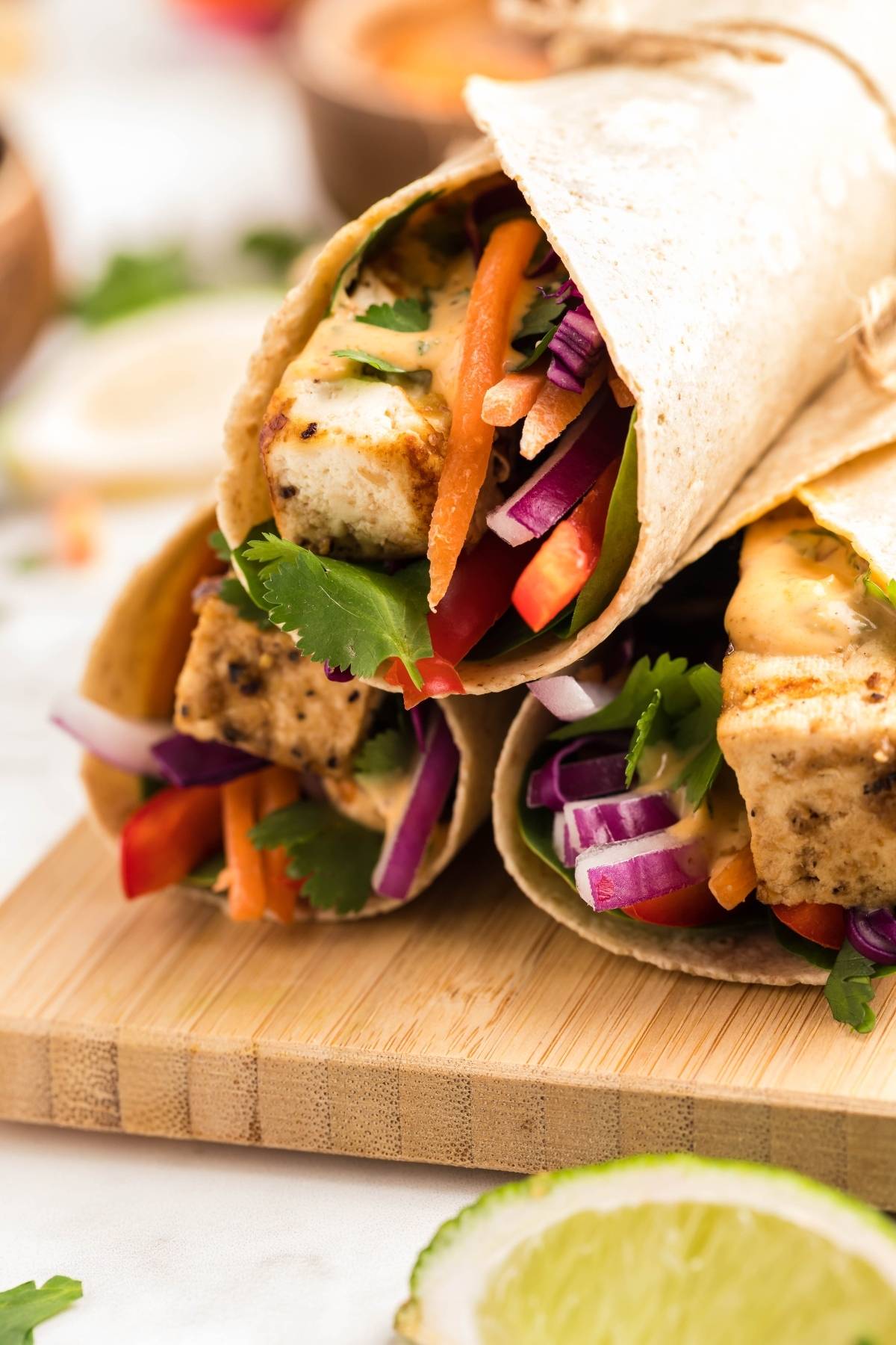 Three Tofu Thai Wraps filled with tofu, red onions, carrots, cilantro, and sauce on a wooden board—perfect for a vegan gluten free lunch. Pair with our Coconut Almond Butter Dip for extra flavor!.