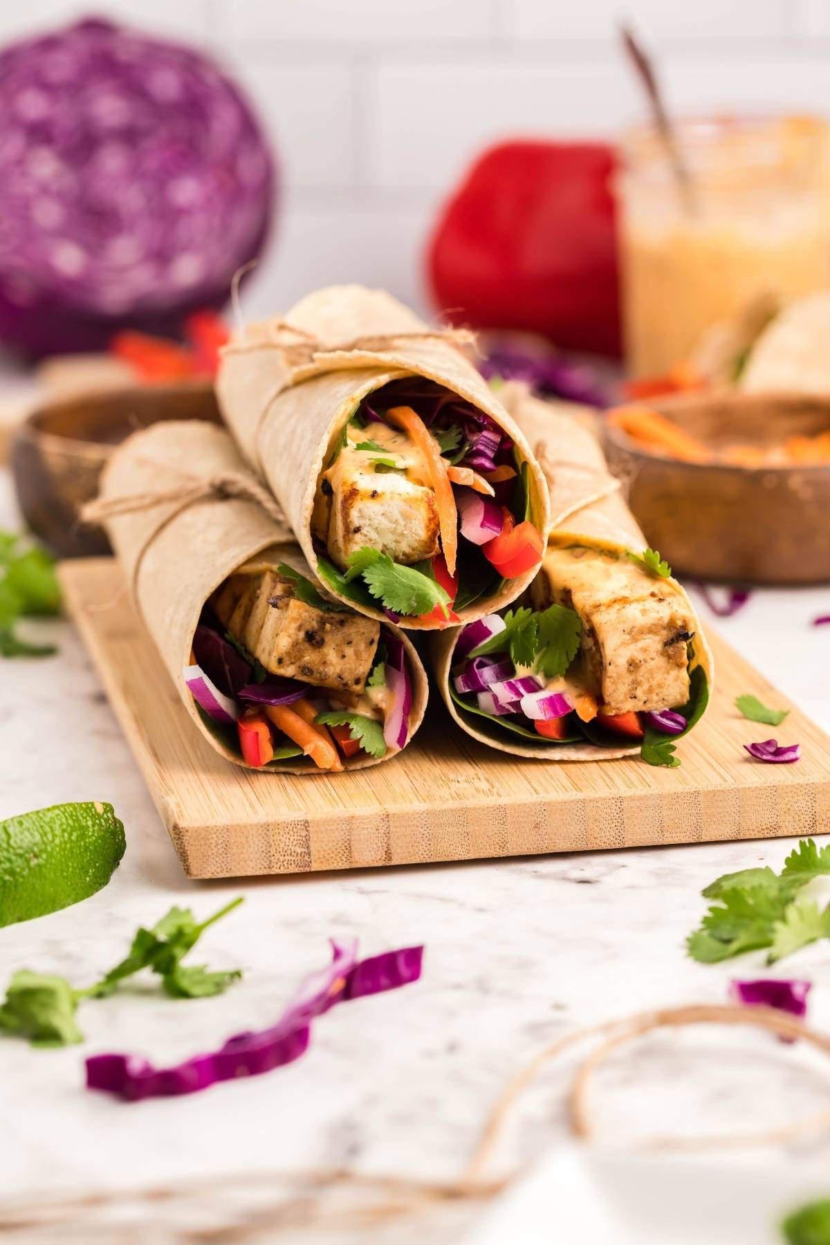 Three easy tofu wraps with red cabbage and cilantro on a wooden board, surrounded by fresh ingredients. Perfect as a vegan and gluten free wrap, they pair deliciously with our creamy Coconut Almond Butter Dip.