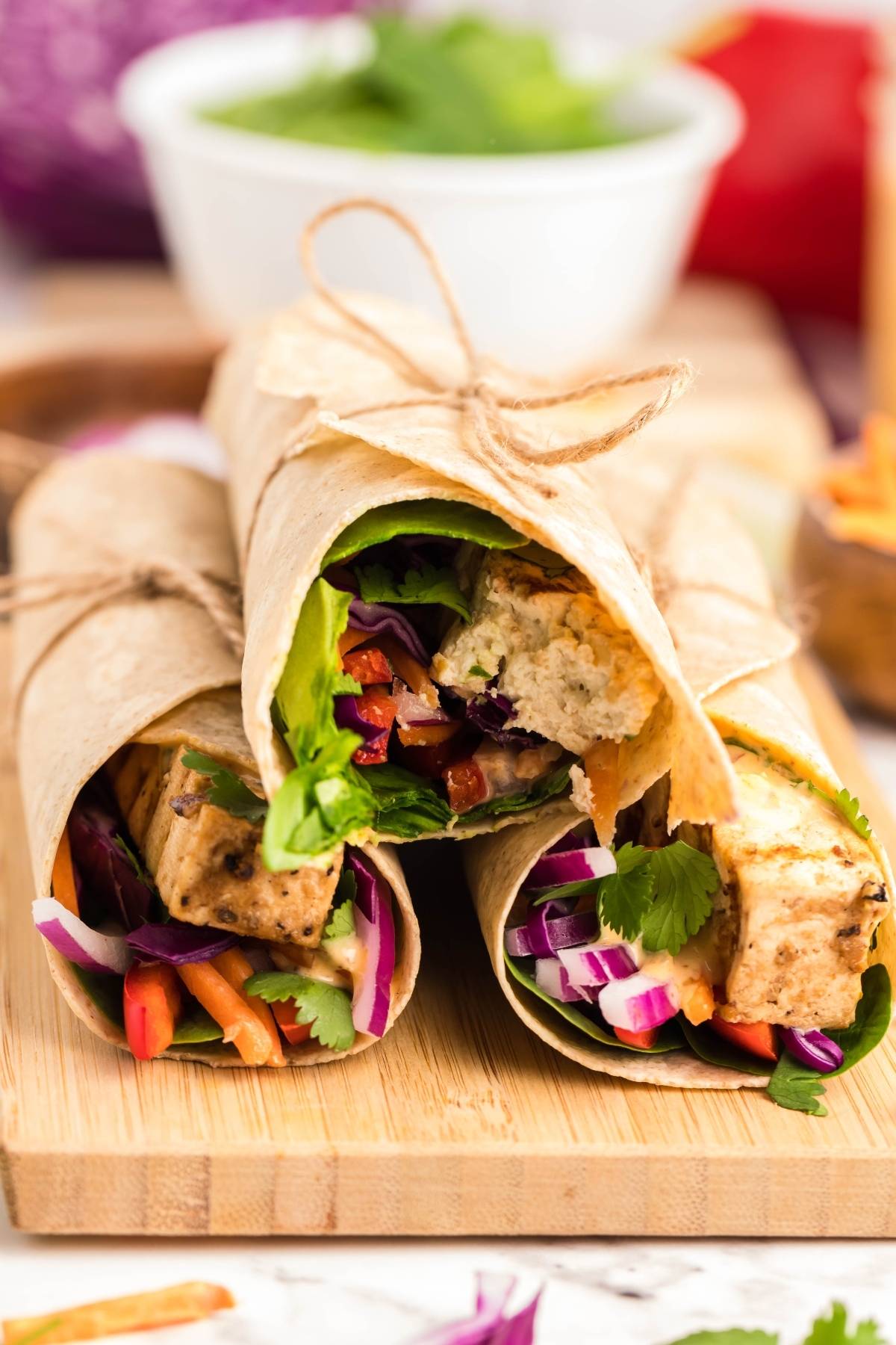 Three vegan and gluten free wraps with tofu, colorful vegetables, and greens, tied with string on a wooden board—perfect as a Tofu Thai Wrap paired with Coconut Almond Butter Dip.