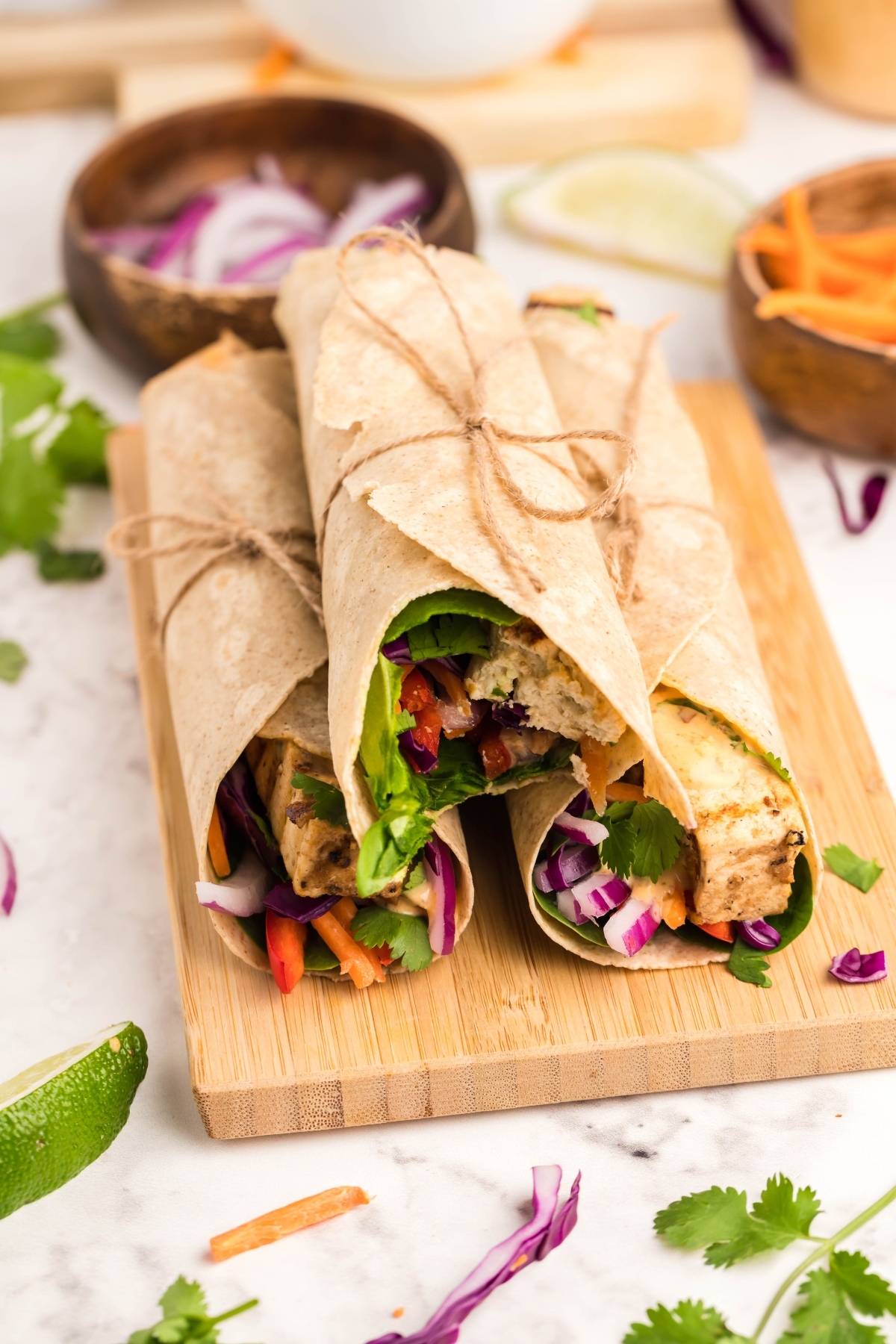 Three vegan and gluten free wraps tied with twine on a wooden board, surrounded by fresh herbs and lime wedges—perfect for a light Vegan Gluten free lunch.
