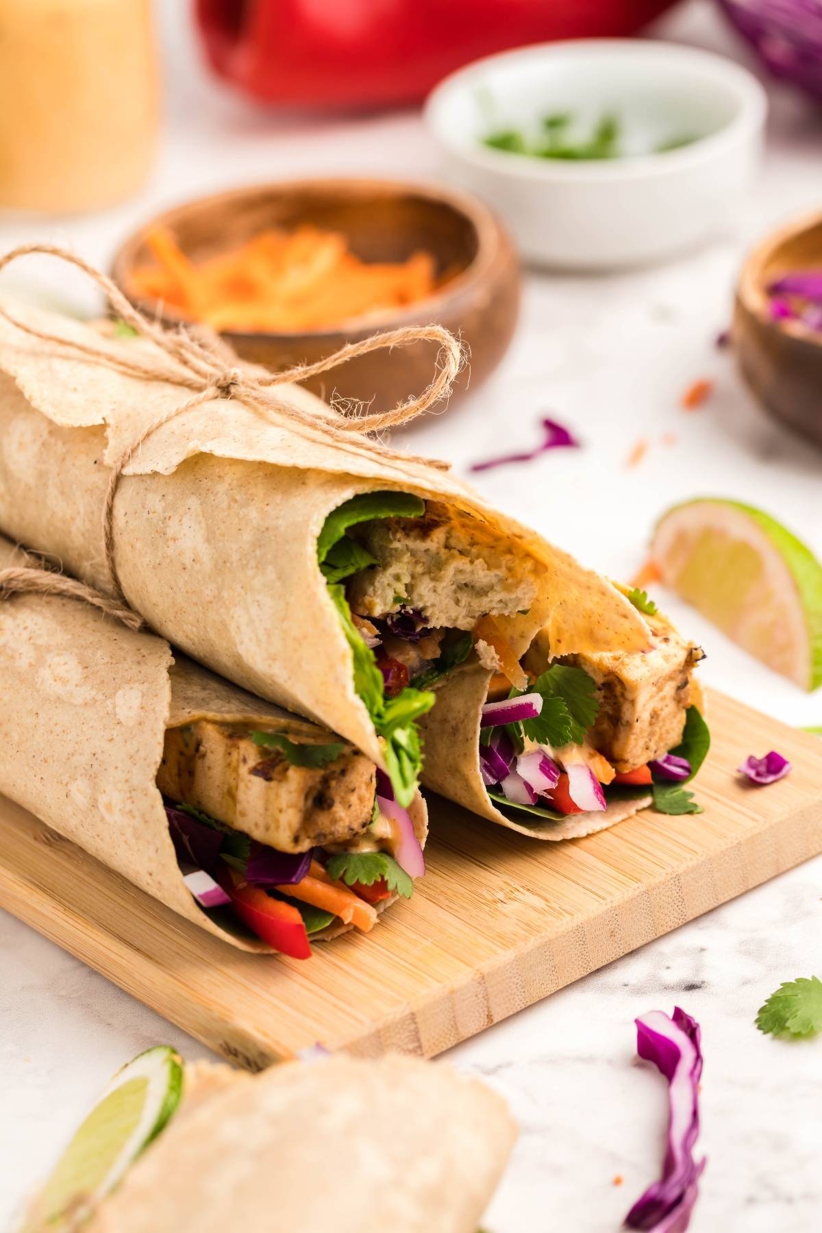 Two Tofu Thai Wraps filled with colorful vegetables and herbs, tied with twine, rest on a wooden board. This vegan and gluten free wrap is perfect for a vibrant, healthy meal.