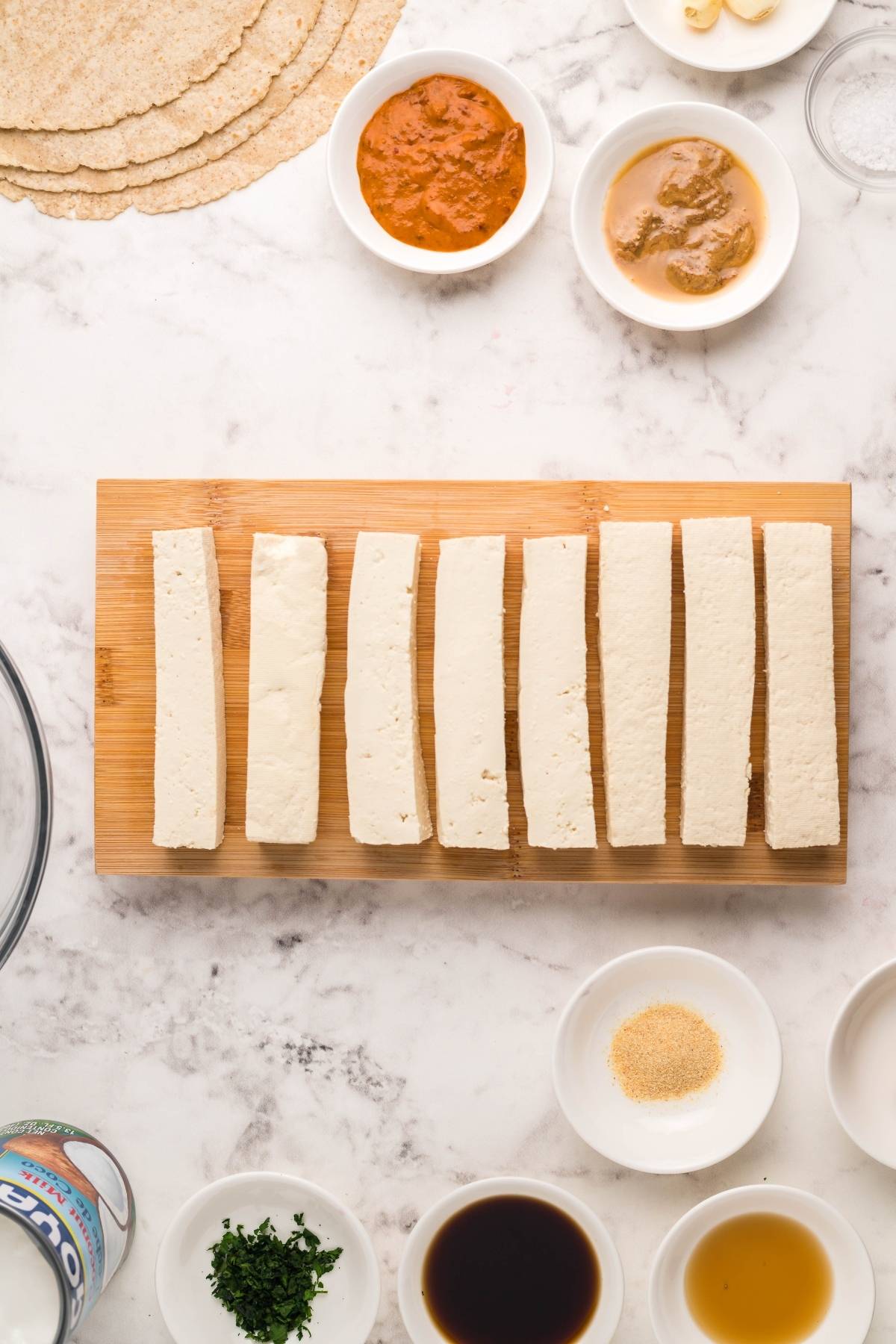 Sliced tofu on a wooden board, surrounded by small bowls of sauces, seasonings, and tortilla wraps—perfect for assembling a Vegan and gluten free wrap or a flavorful Tofu Thai Wrap with Coconut Almond Butter Dip.