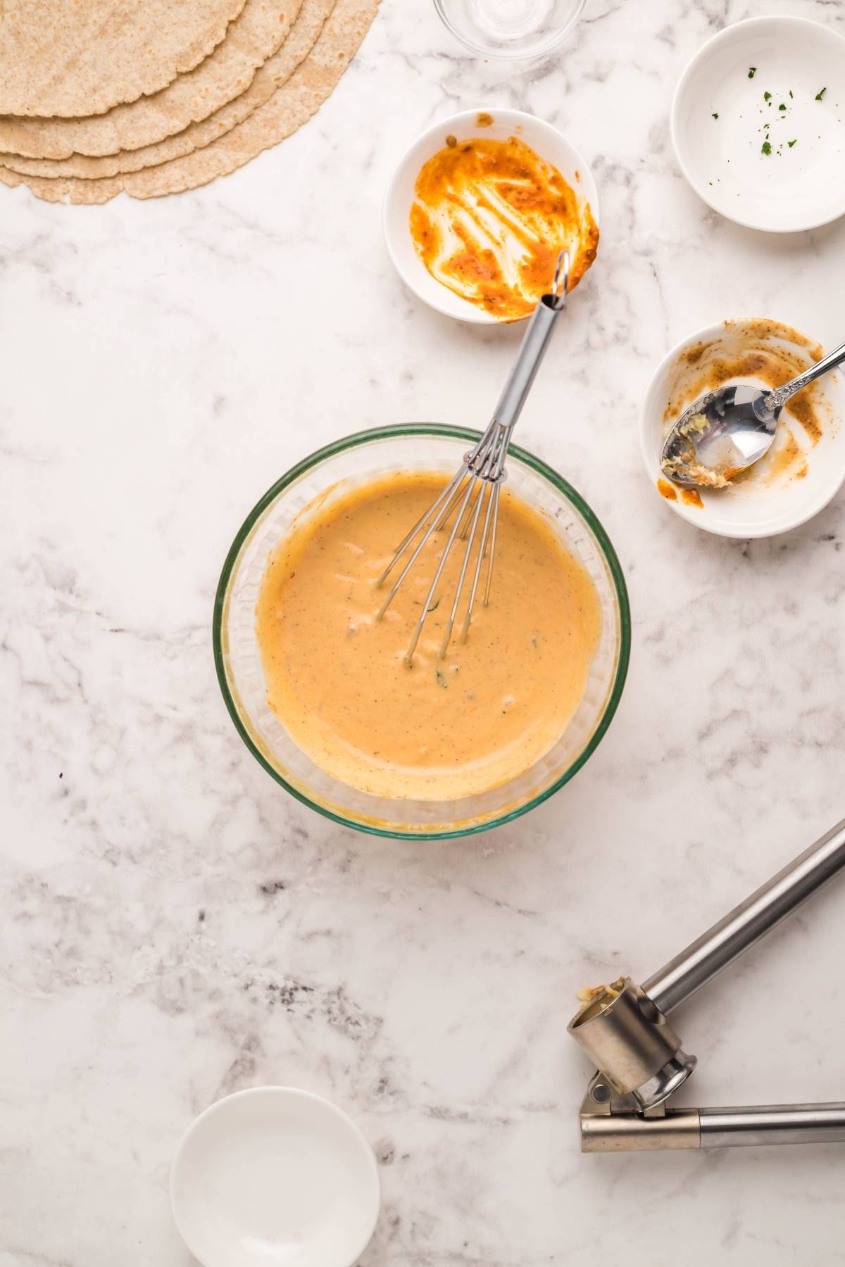 A bowl of orange Coconut Almond Butter Dip with a whisk, surrounded by dirty bowls, utensils, and tortillas for a Vegan and gluten free wrap on a marble counter.