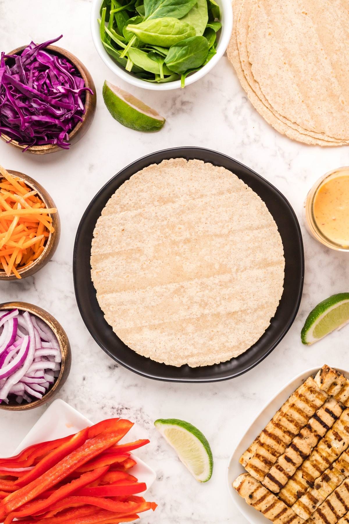 A Vegan and gluten free wrap: a tortilla on a plate surrounded by vegetables, grilled tofu, lime wedges, and sauce on a marble surface.