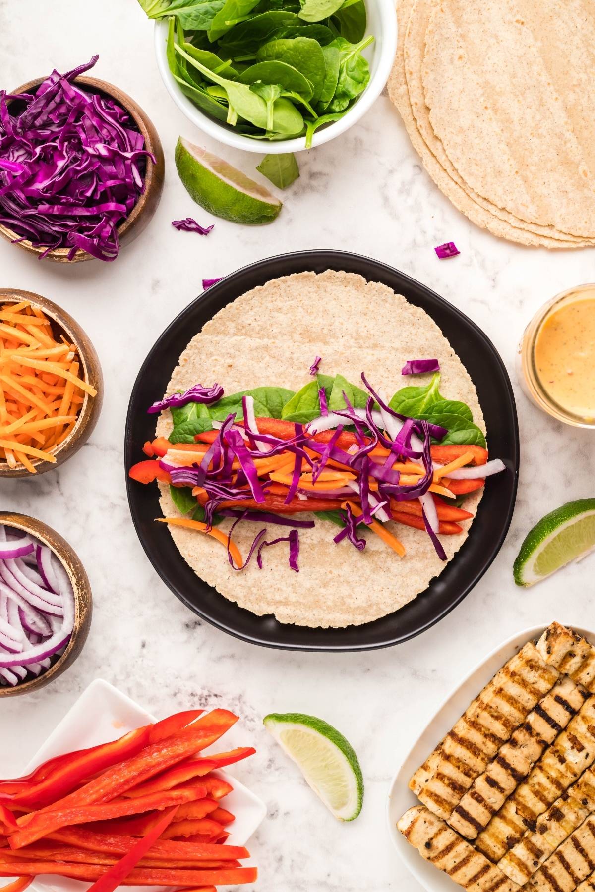 A vegan and gluten free wrap topped with spinach, red cabbage, carrot, and sauce, surrounded by fresh veggies and grilled tofu—perfect for a delicious vegan gluten free lunch.