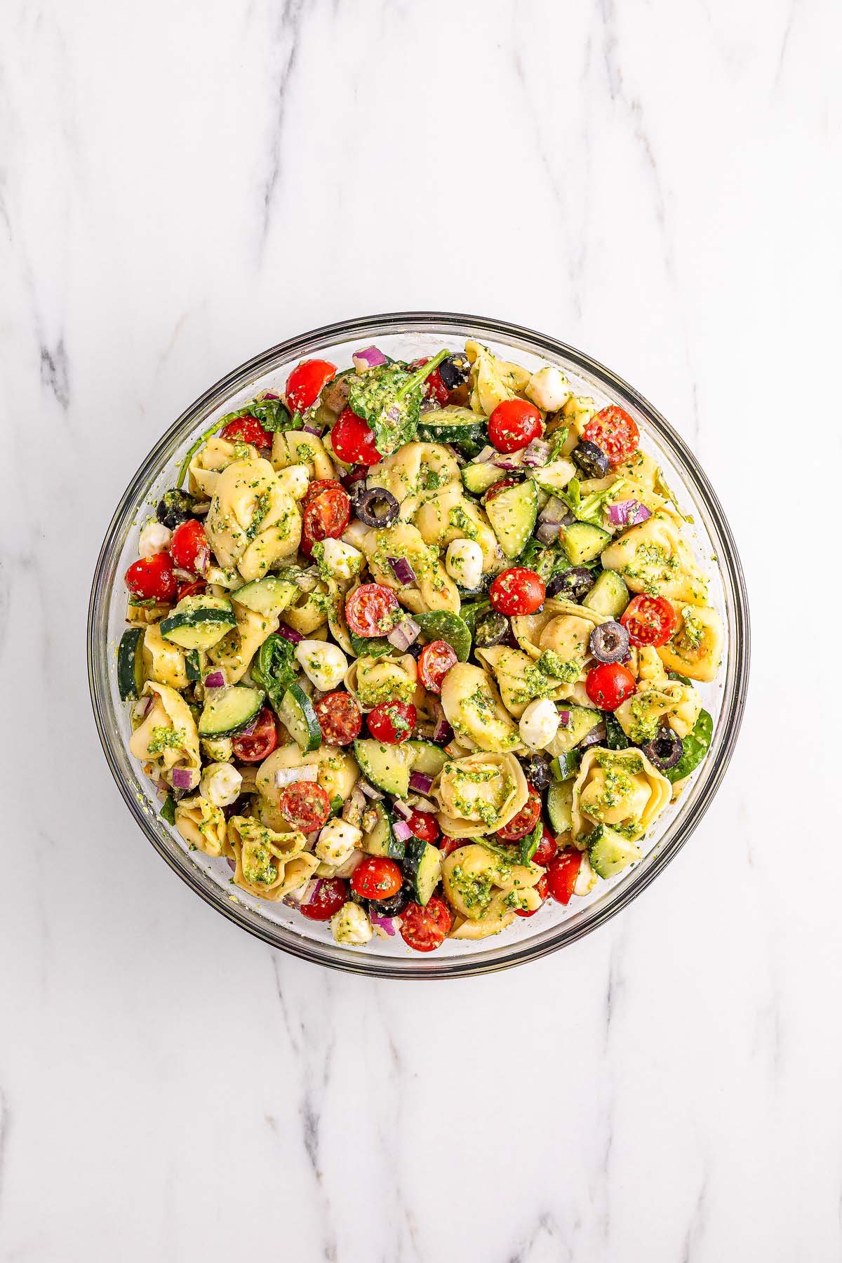 Bowl of pesto tortellini pasta salad with tomatoes, cucumbers, olives, mozzarella, and herbs on a white surface.