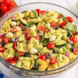 A bowl of pesto tortellini pasta salad with mozzarella, tomatoes, cucumbers, and olives sits on a white table.