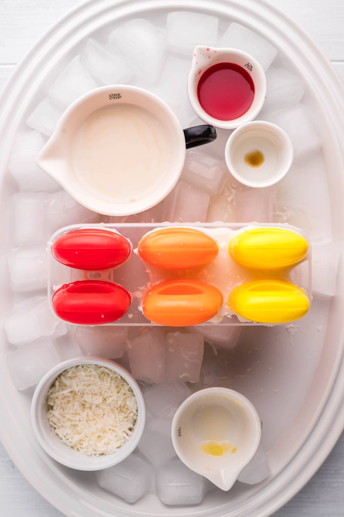 Colorful popsicle molds with bowls of milk, red liquid, vanilla, shredded coconut, and ice cubes on a tray—perfect for making a layered popsicle recipe or delicious coconut ice pops.