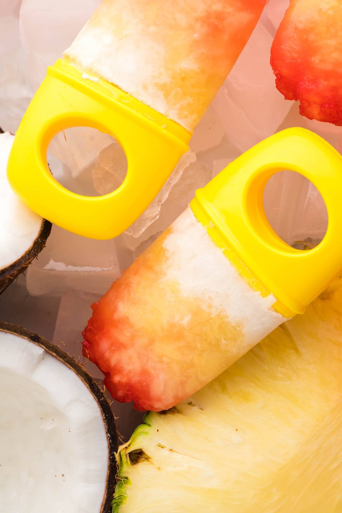Two colorful Tropical Sunrise Popsicles with yellow handles rest on ice, surrounded by coconut and pineapple slices—perfect for trying a coconut ice pops or layered popsicle recipe at home.