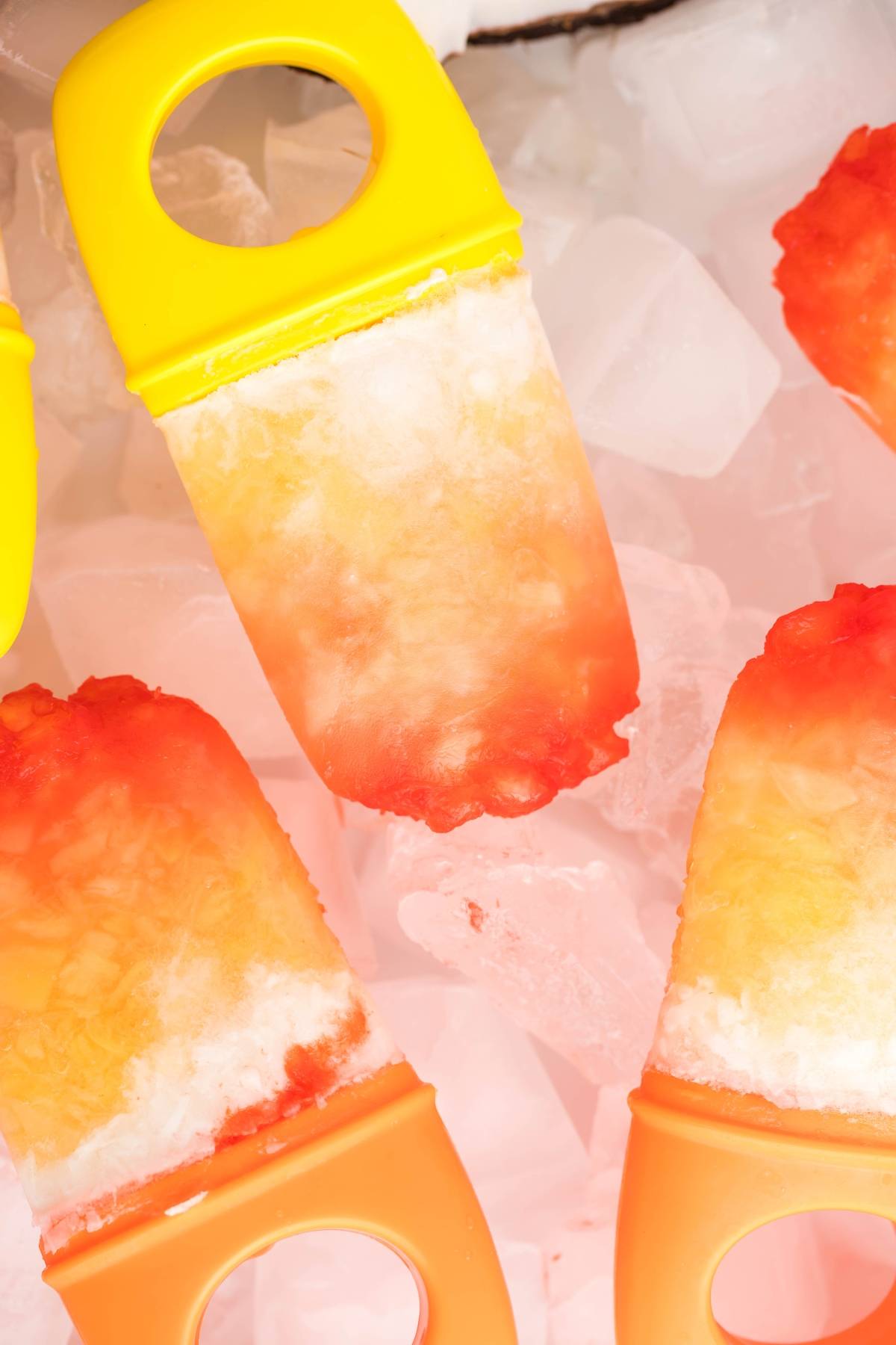 Colorful homemade popsicles with yellow and orange handles rest on a bed of ice cubes, showcasing a delicious layered popsicle recipe that's perfect for making coconut ice pops at home.
