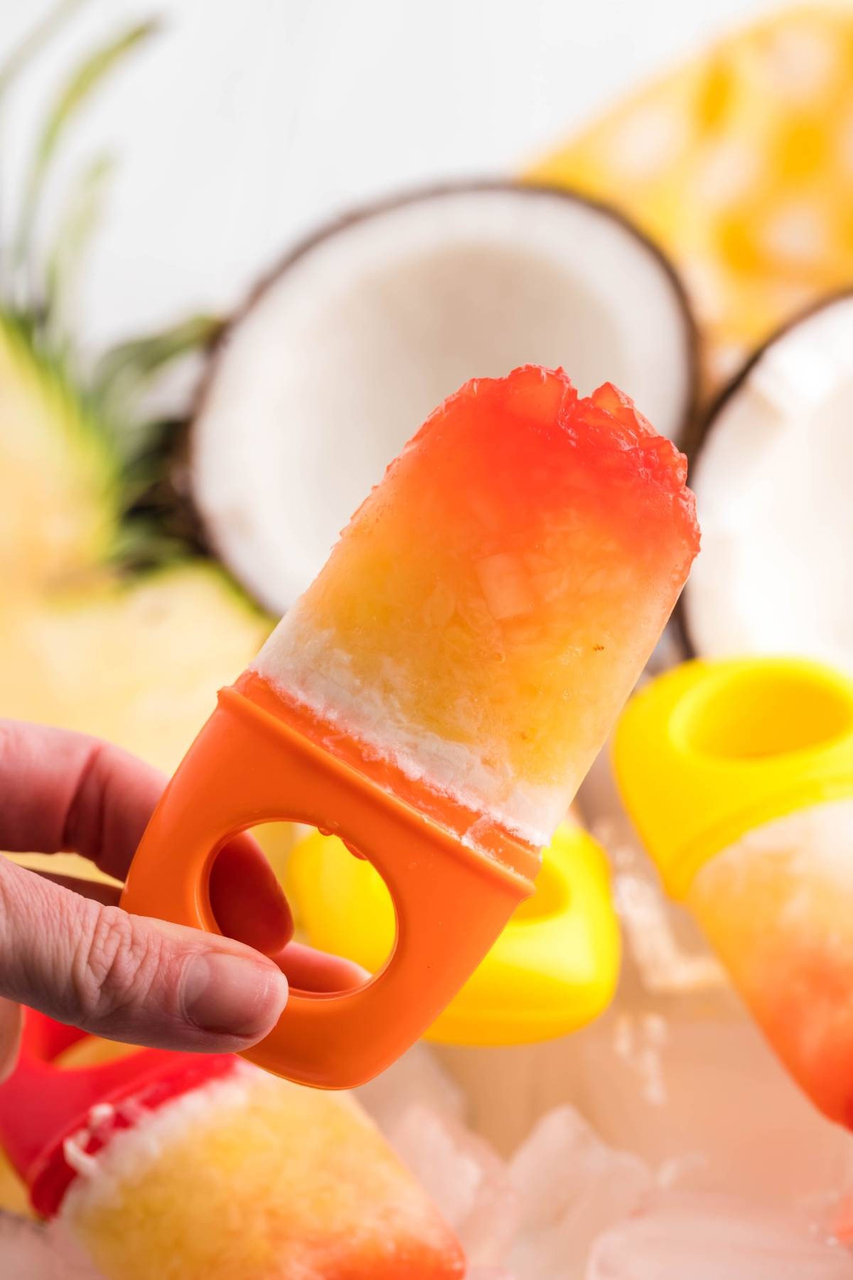 A hand holds a colorful Tropical Sunrise Popsicle with orange, yellow, and white sections in a plastic mold—a perfect layered popsicle recipe for summer treats.