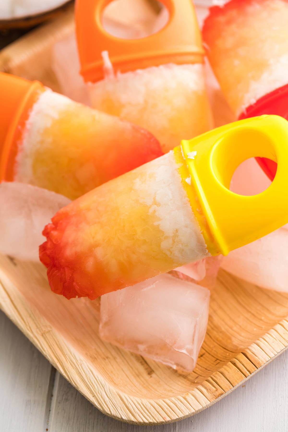 Colorful homemade Tropical Sunrise Popsicles with orange and yellow handles rest on a wooden tray with ice cubes, showcasing a layered popsicle recipe perfect for summer.