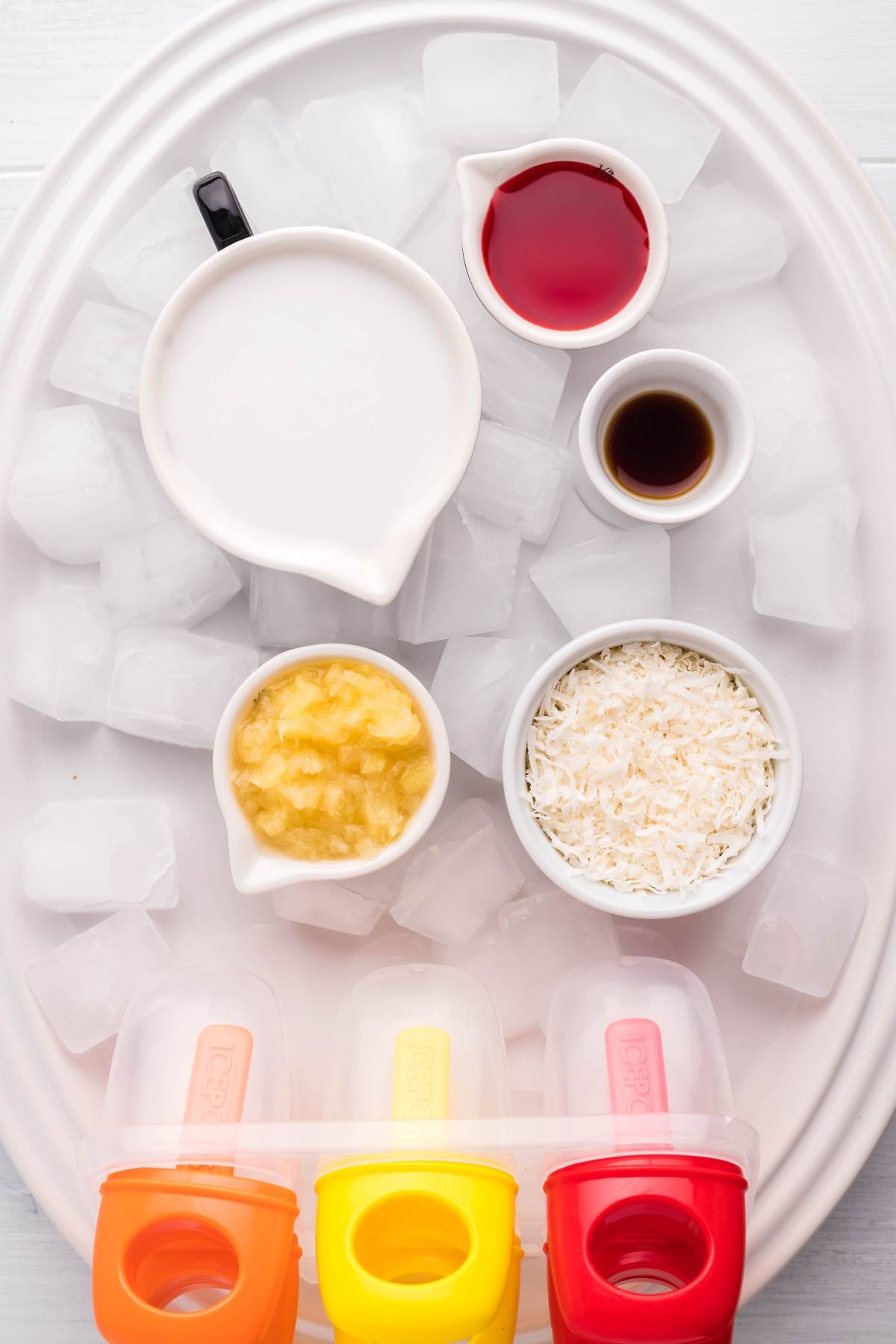 Five ingredients in small bowls on ice (coconut milk, juice, vanilla, pineapple, coconut) with empty popsicle molds—perfect for creating Tropical Sunrise Popsicles or trying a fun layered popsicle recipe at home.
